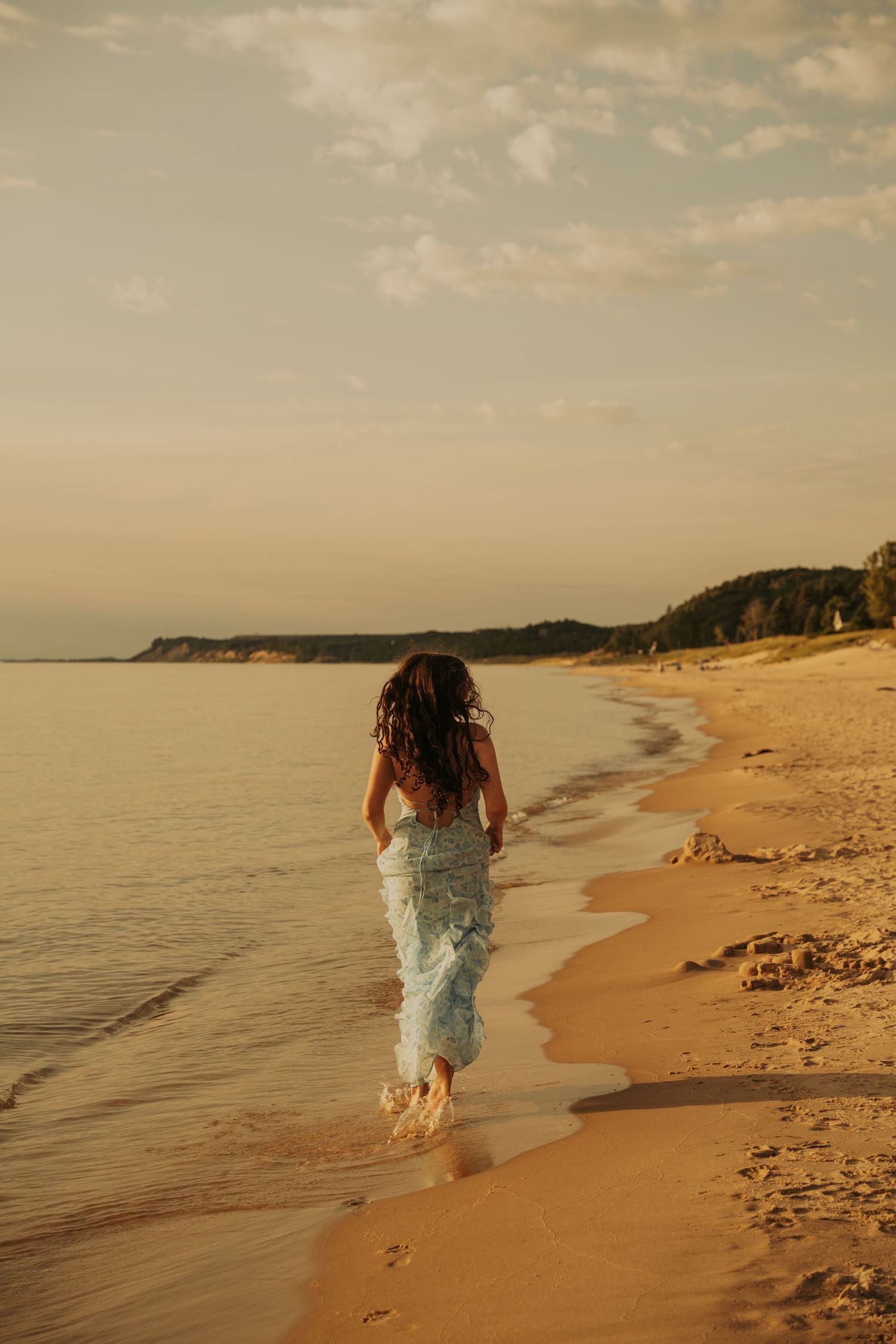 A person in a flowy dress walks along a serene beach at sunset with warm golden light illuminating the coastline.
