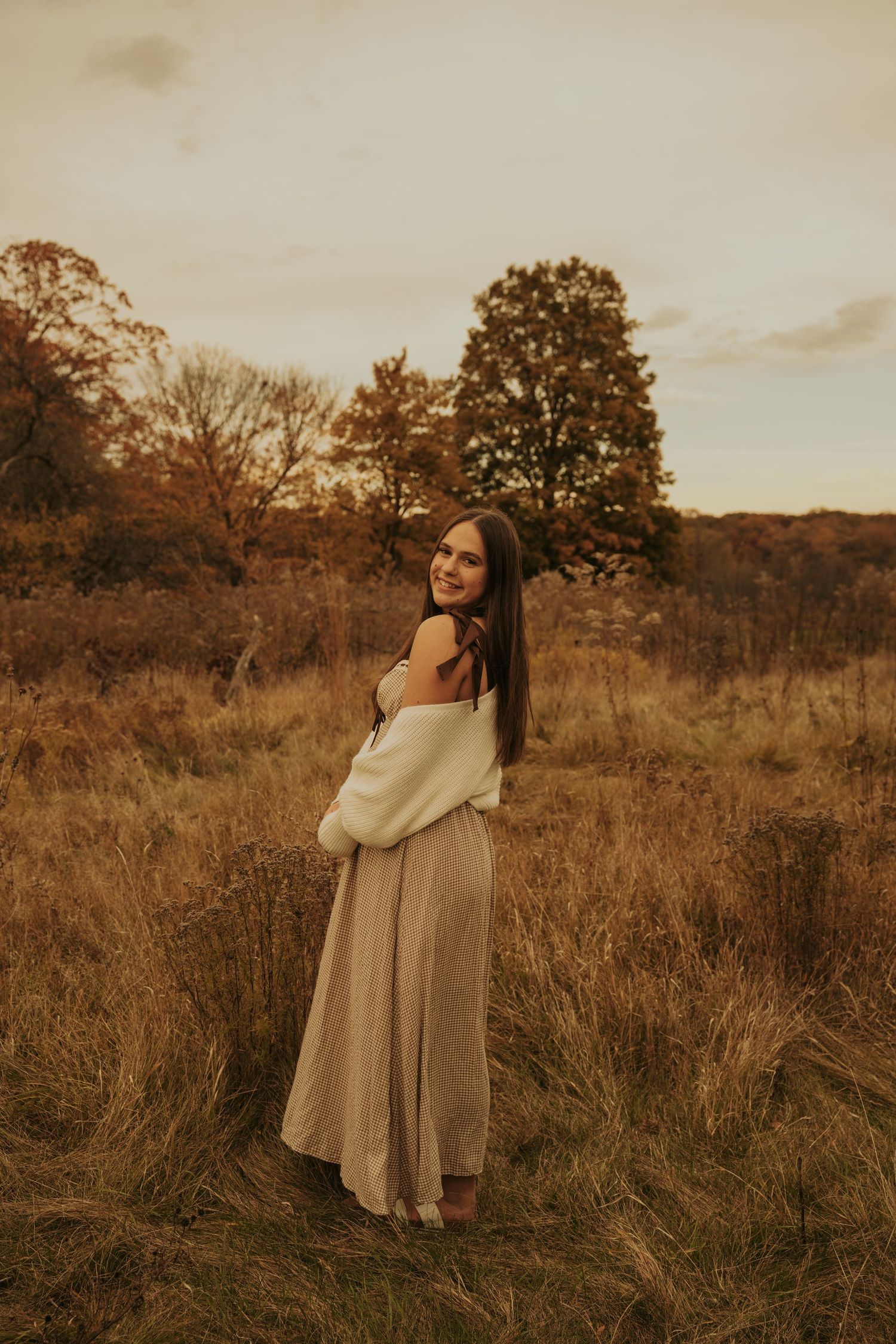 A couple embraces in a golden autumn field at sunset with tall grass and trees in the background.