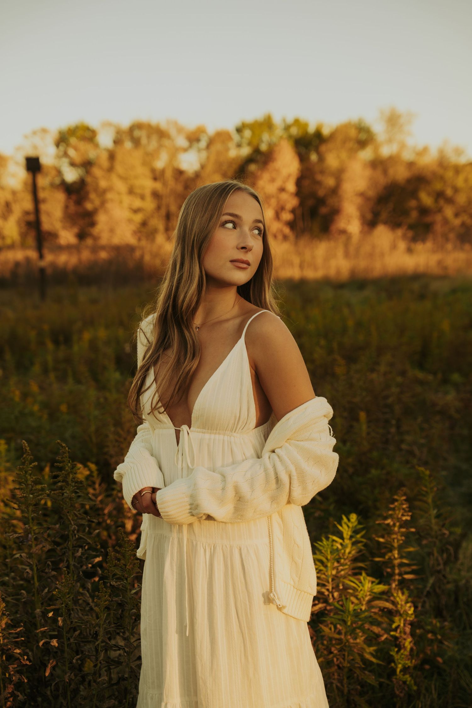 A person in a flowing white dress and cardigan poses in a field at golden hour with autumn trees in the background.