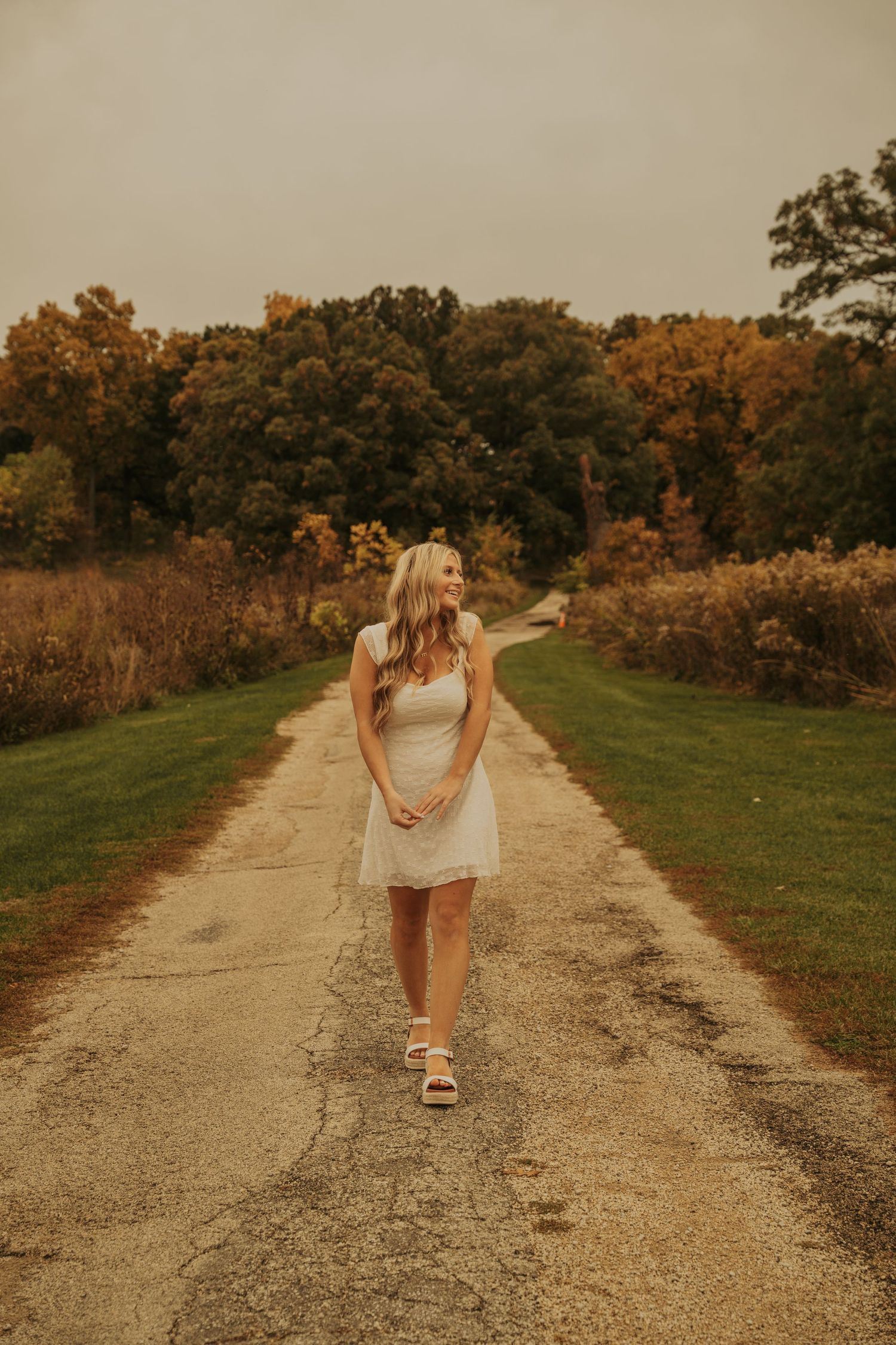 A person in a white dress walks down a gravel path surrounded by autumn trees during golden hour.