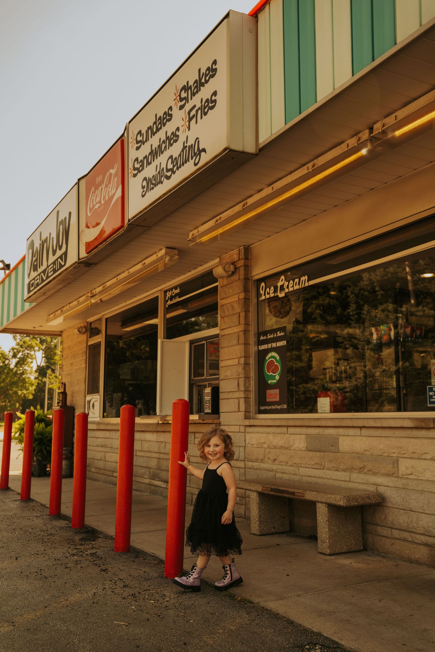 A row of red bollards line the front of a strip mall storefront with green trim and wooden benches at sunset.