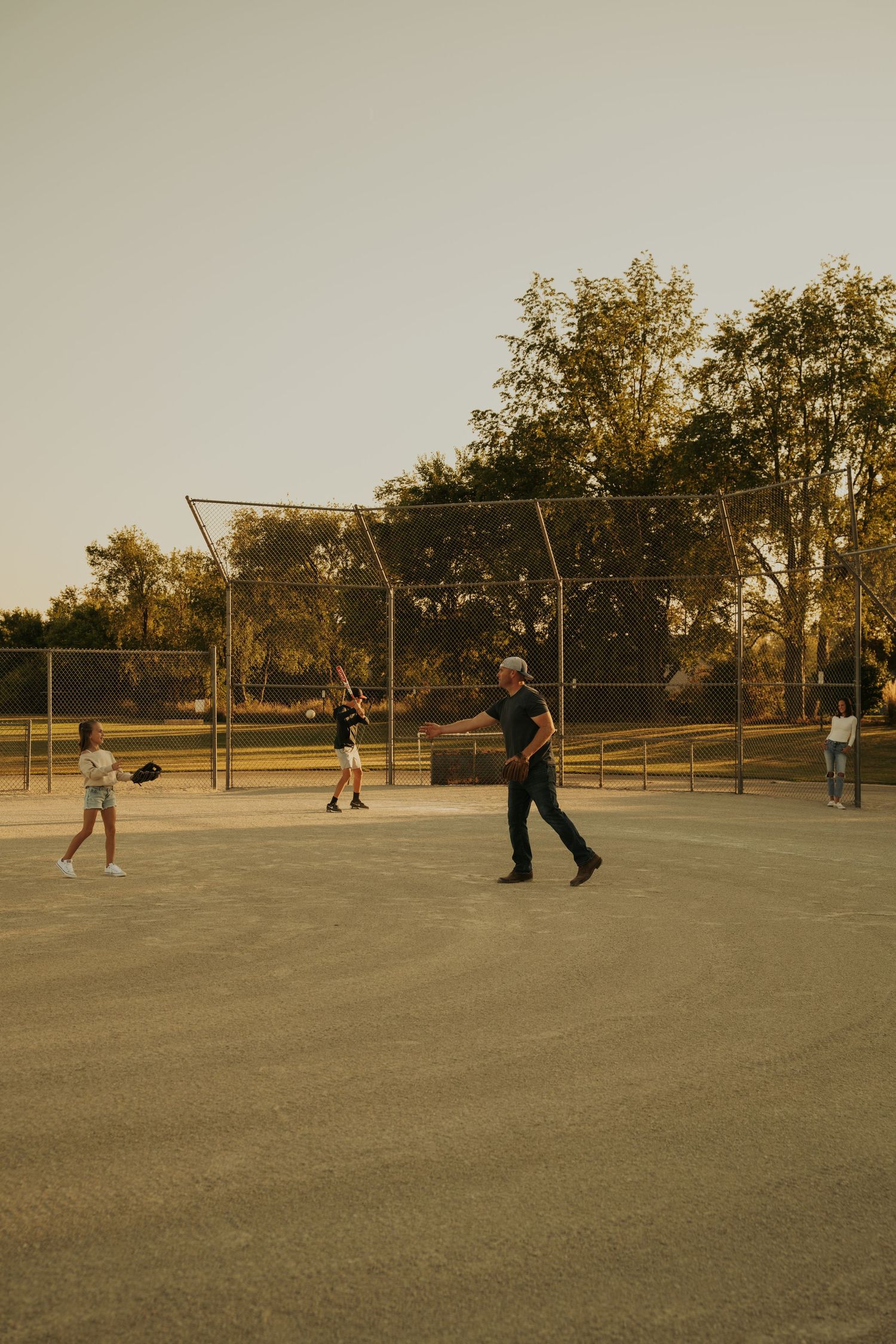 People playing baseball on a dusty field at sunset with trees visible in the background.