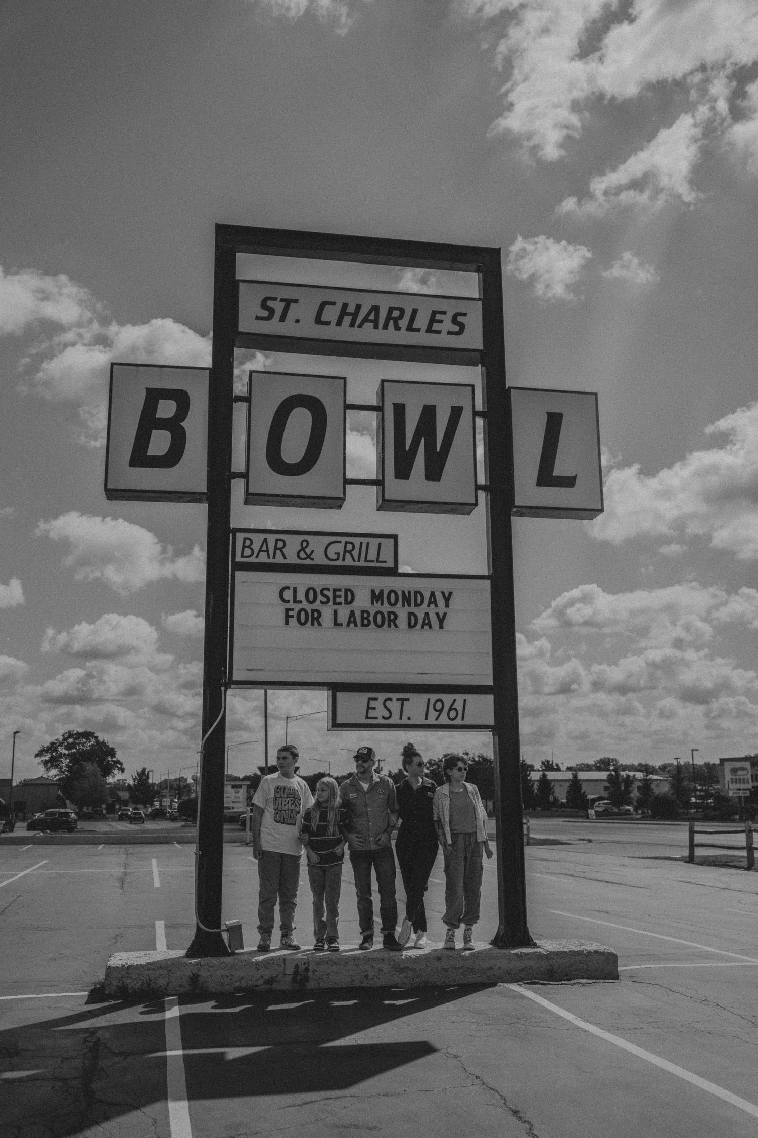 Black and white photo of people standing beneath vintage St. Charles Bowl bar and grill sign with cloudy sky background.