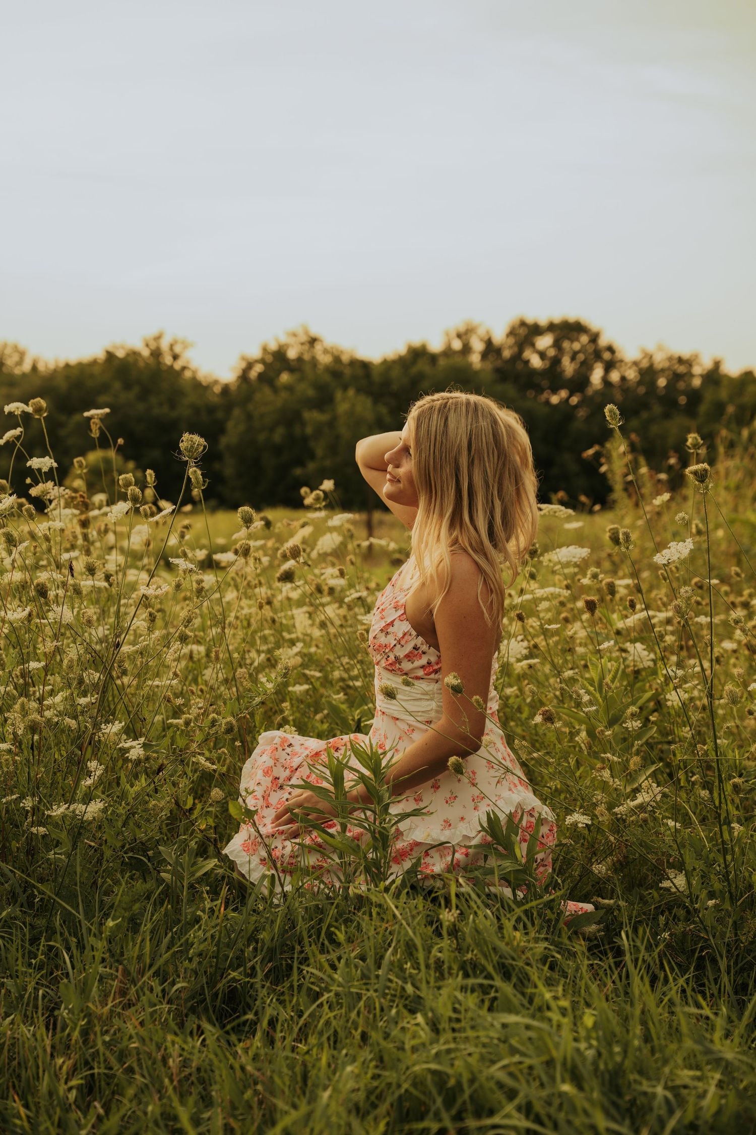 Someone in a floral dress sits among wildflowers in a golden summer meadow at sunset.
