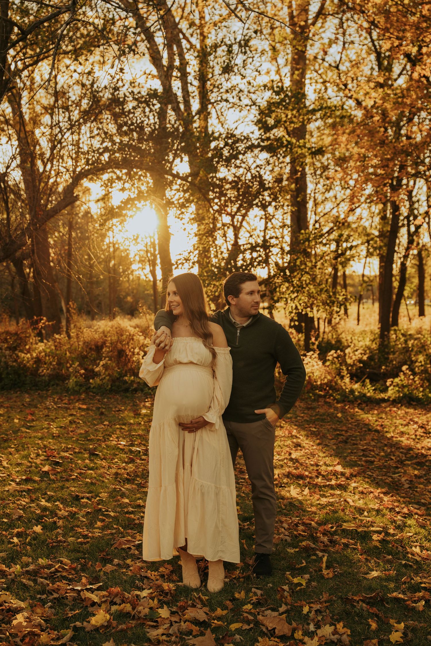 A couple stands in a golden autumn forest at sunset with fallen leaves scattered on the ground.