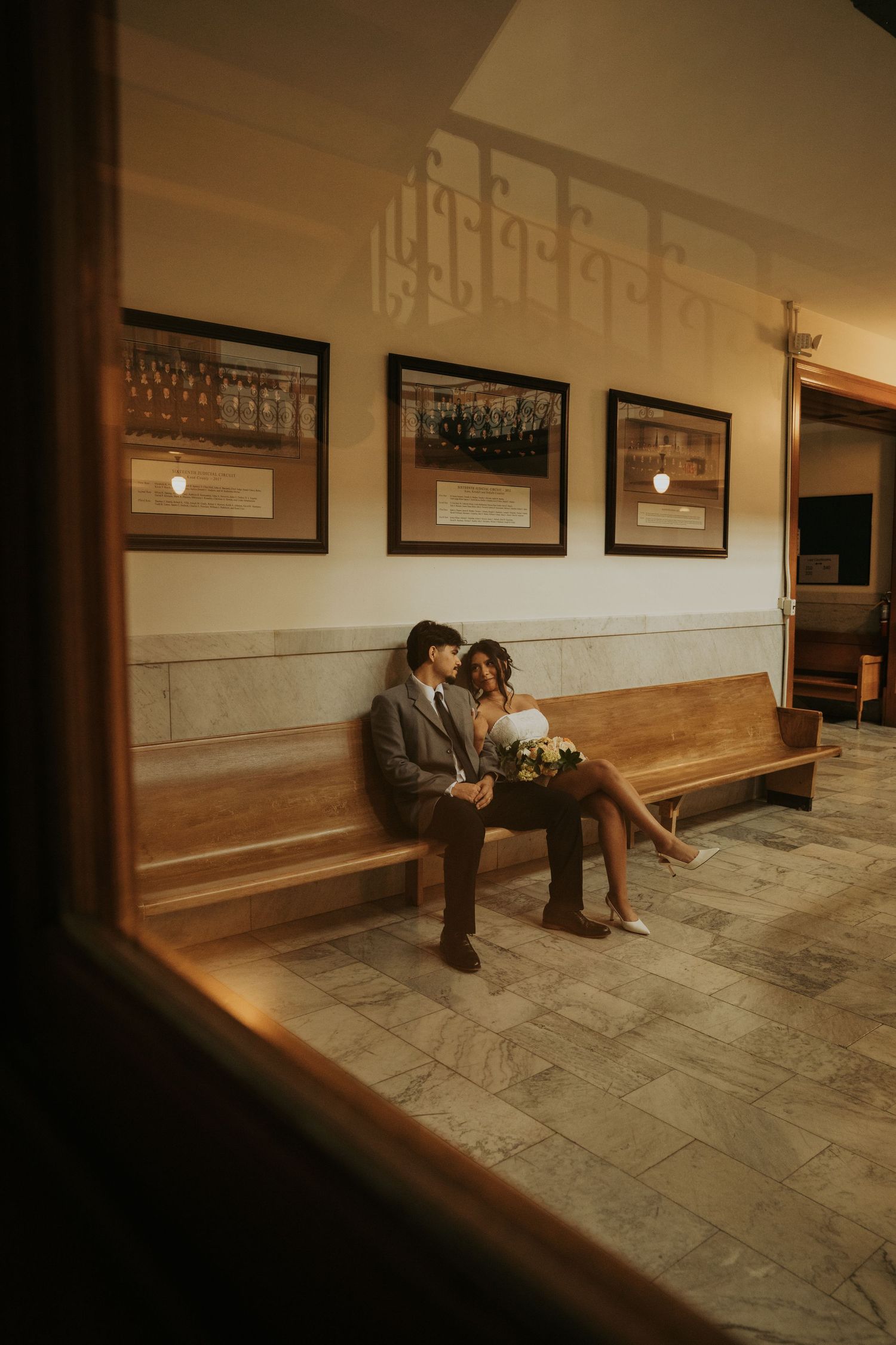 Two people sit together on a wooden bench in a dimly lit hallway with framed artwork on the wall.
