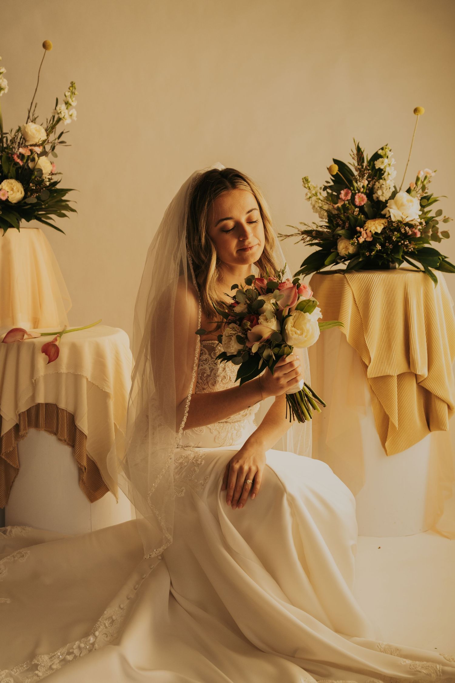 A bride in a white wedding dress holds a floral bouquet while seated among elegant table arrangements with yellow linens.