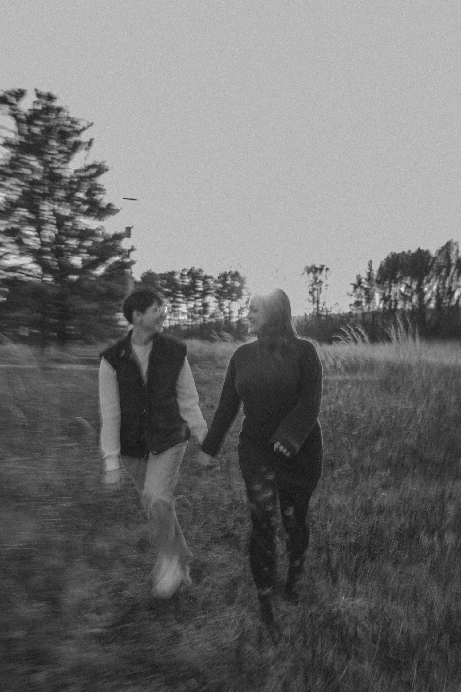 A couple walks hand in hand through a grassy field at dusk in black and white.
