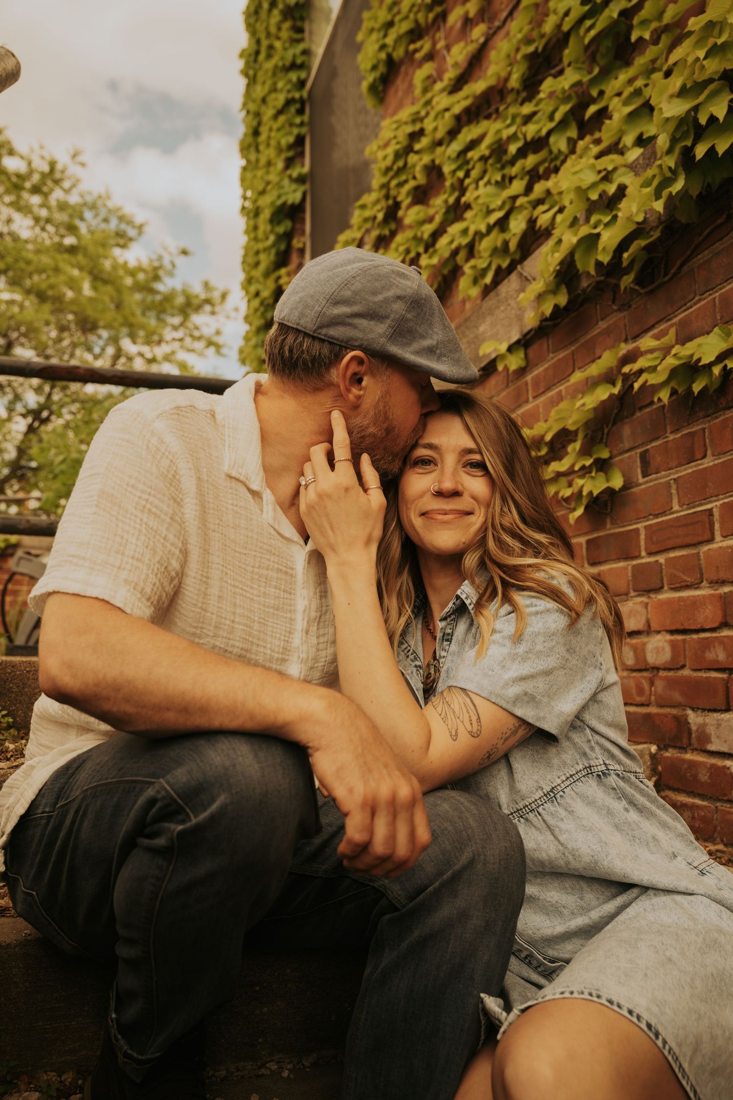 A romantic couple sitting together outdoors against a brick wall with green ivy in warm sunlight.