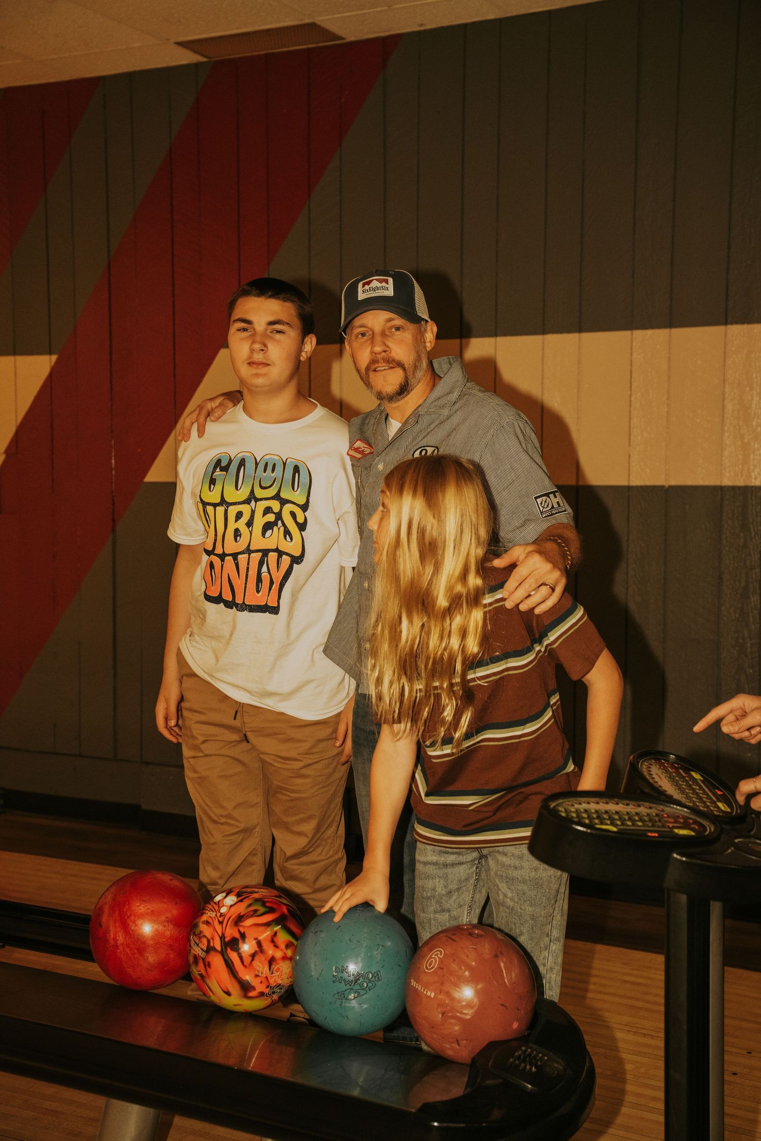 A group of people standing near colorful bowling balls at a bowling alley in warm lighting.