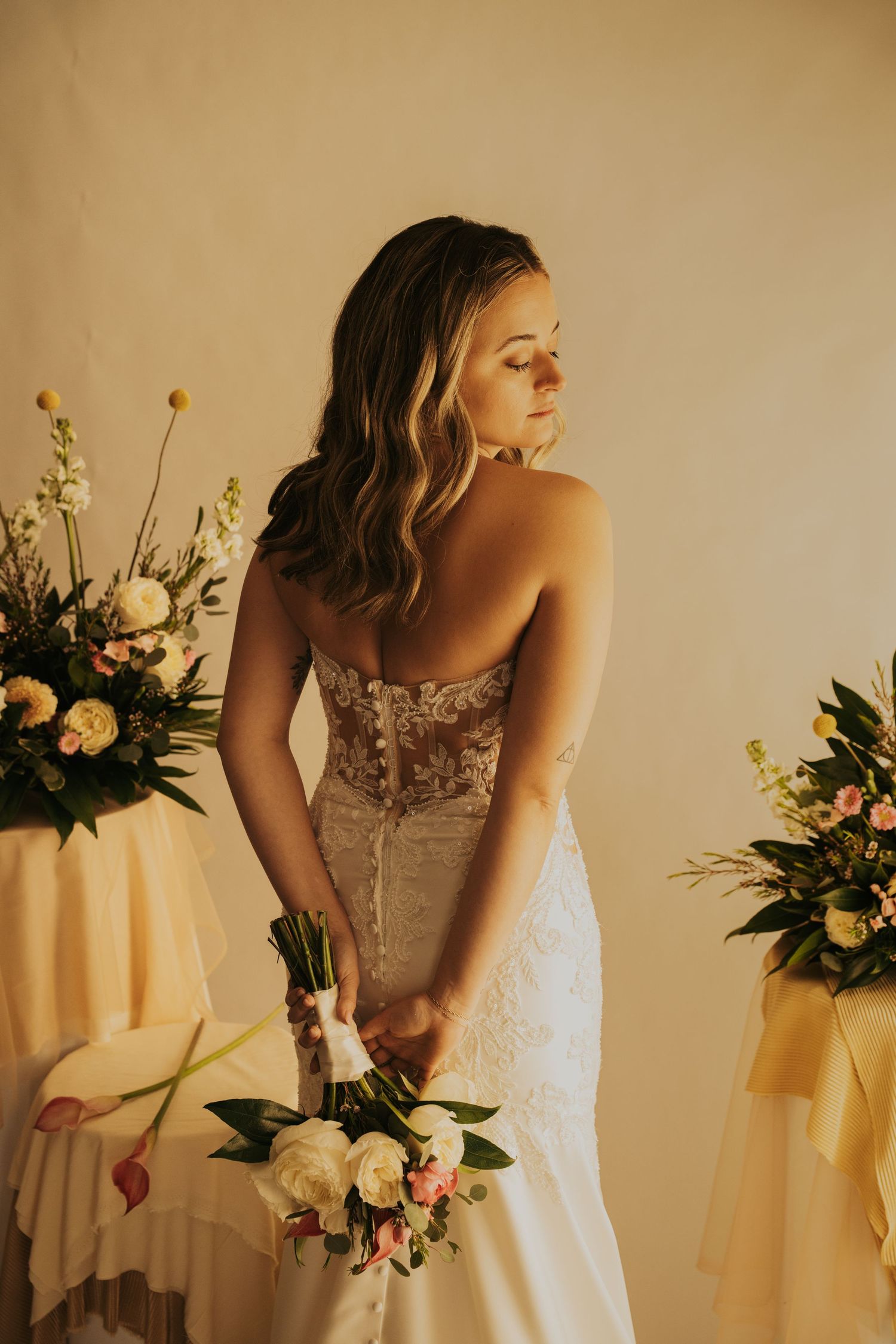 A bride in a lace corset wedding dress holds a bouquet while standing between floral arrangements in warm lighting.
