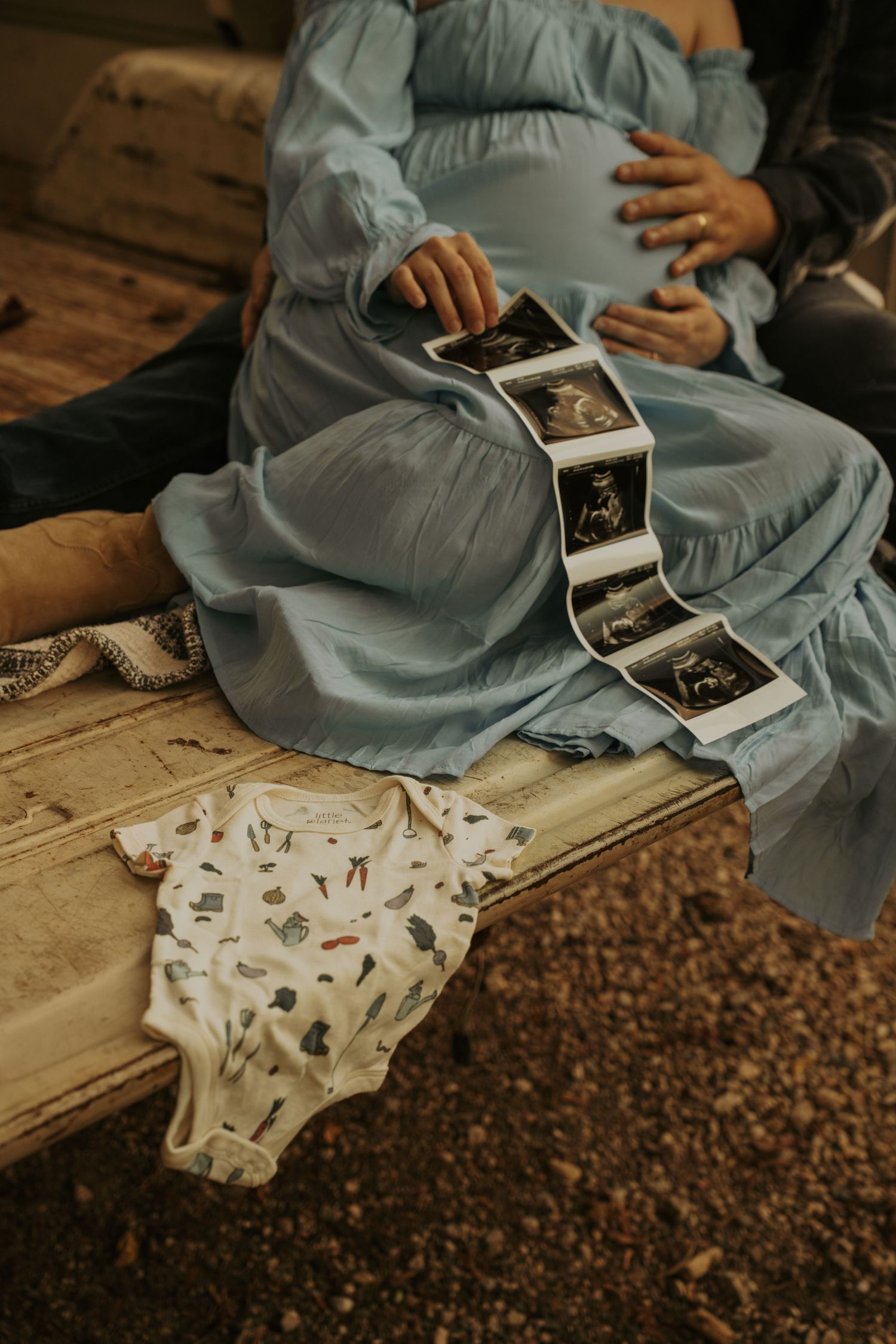 Someone in a blue dress holds ultrasound photos while a baby onesie with small prints lies on wooden surface.