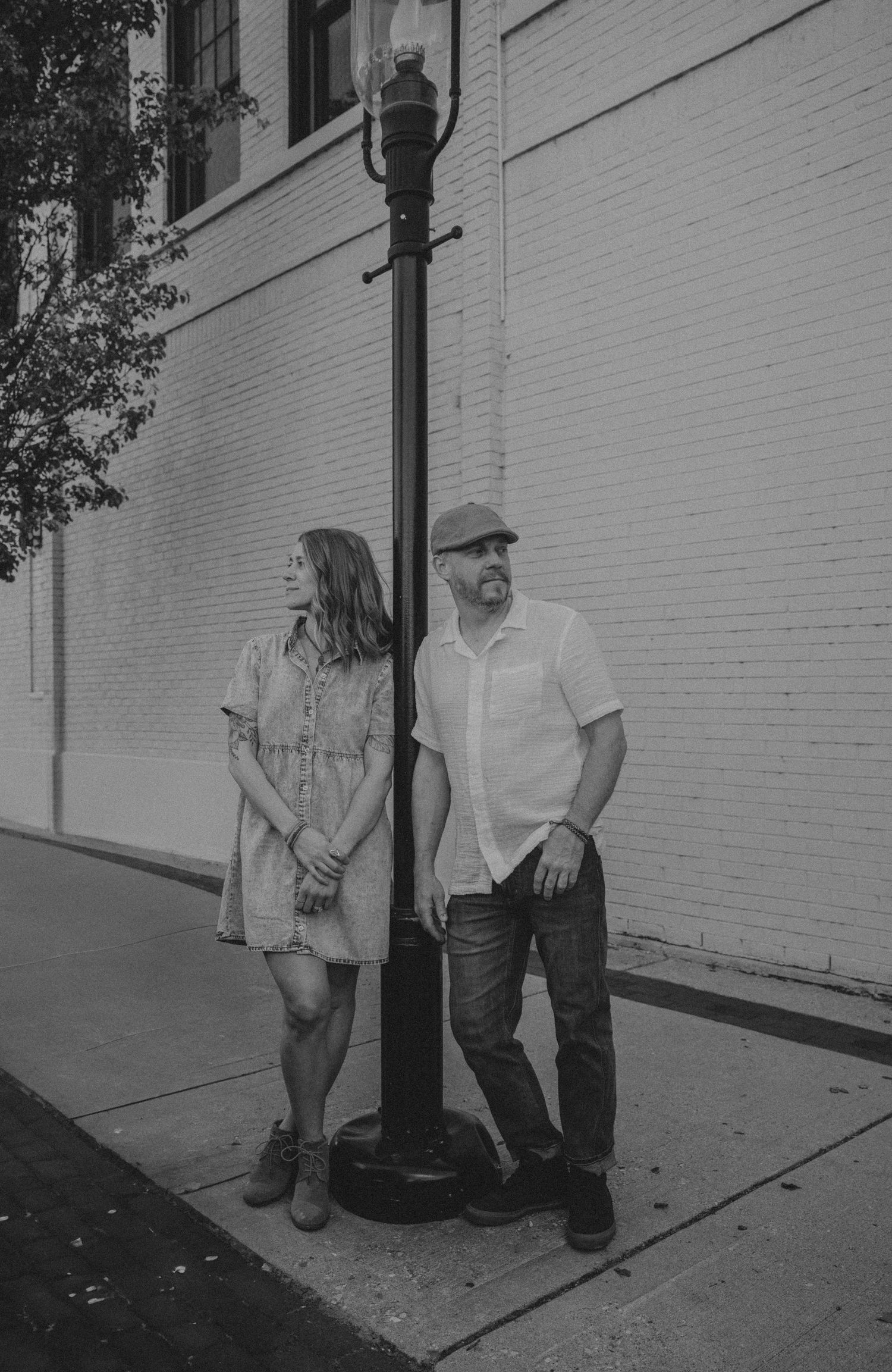 Black and white photo of two people standing by a lamp post against a white brick wall on an urban sidewalk.