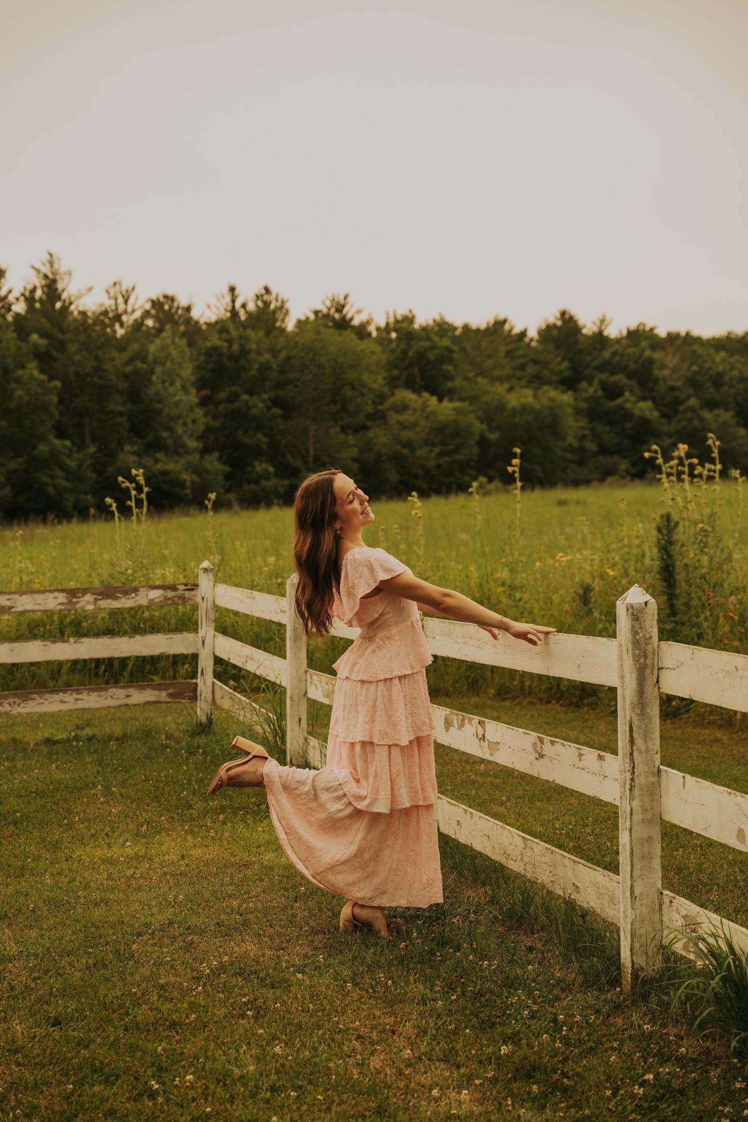 A person in a flowing pink dress dances gracefully along a rustic wooden fence in a sunny meadow field.