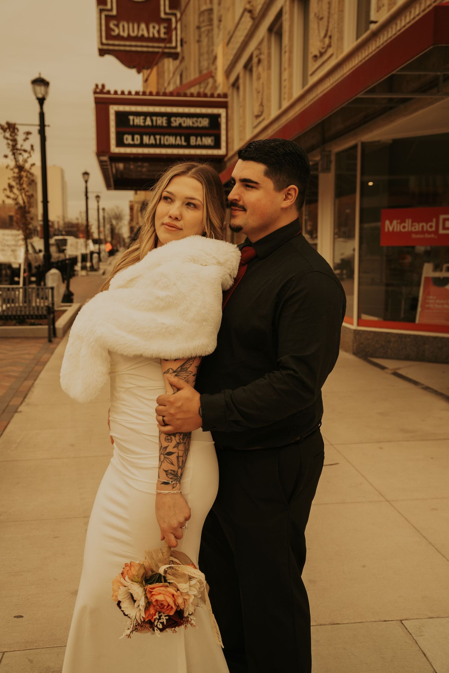 A couple in wedding attire embraces on an empty city sidewalk with vintage storefronts in the background.