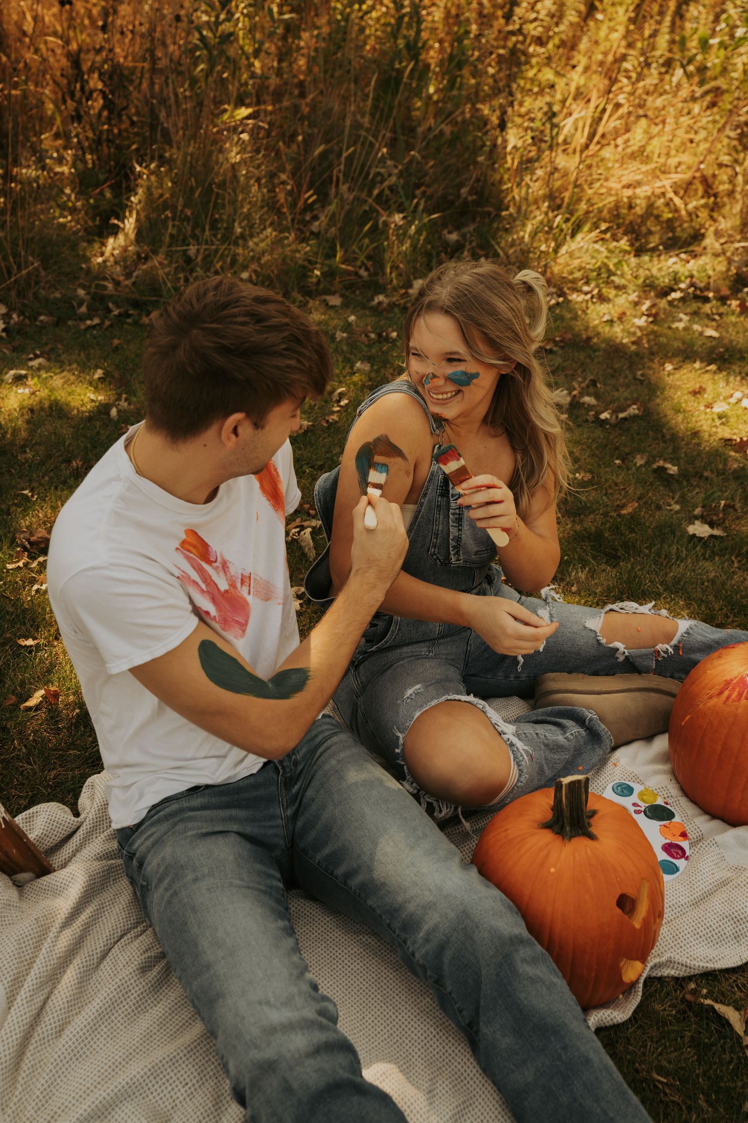 A playful couple paints each other's faces while sitting outdoors with pumpkins on a cozy blanket in autumn lighting.