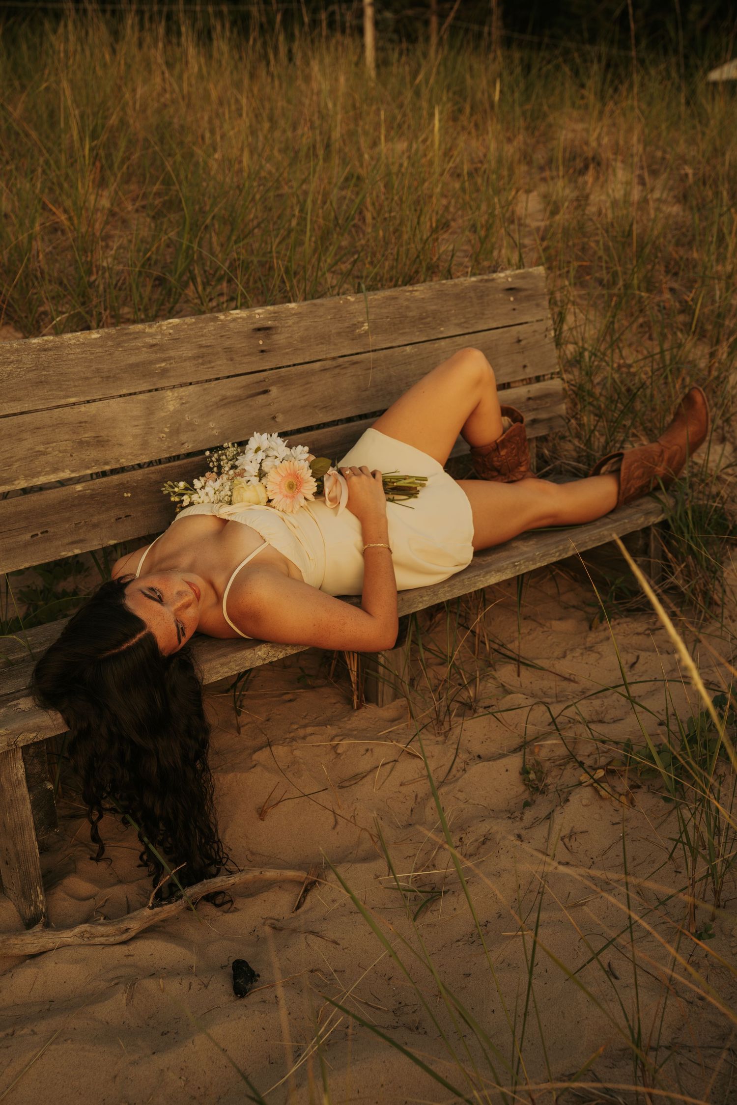 A person in a white dress lies on a wooden bench in a sandy outdoor setting at dusk.
