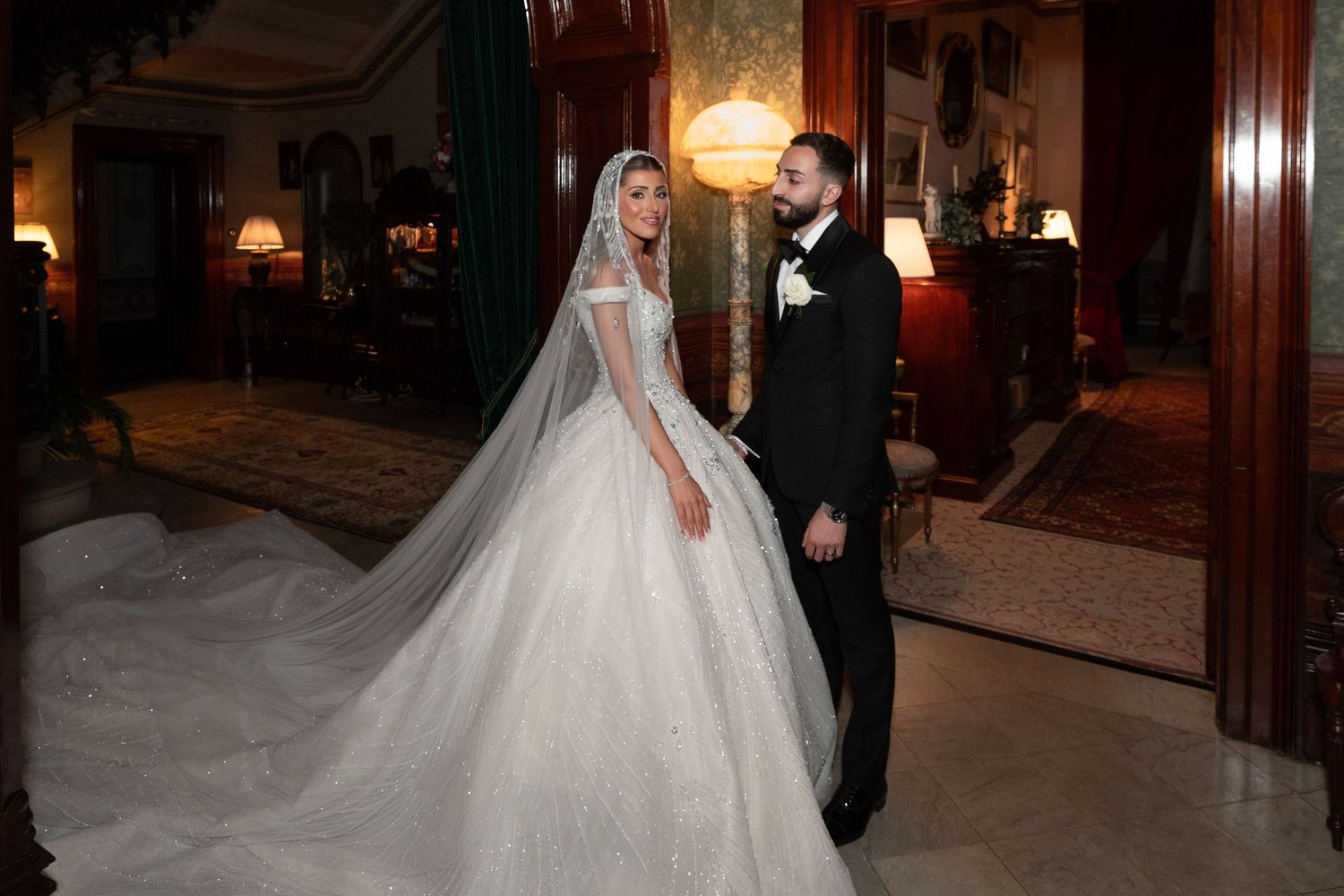 Elegant bride in a long veil and groom in a tuxedo pose in a luxurious room on their wedding day.