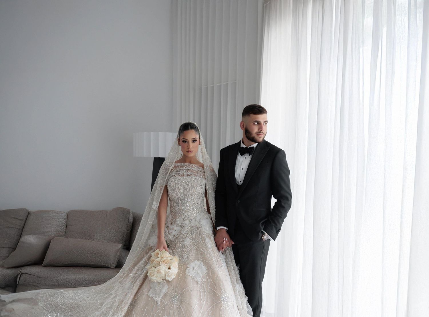 Elegant bride in lace ball gown with veil holds bouquet beside groom in black tuxedo in bright modern room.
