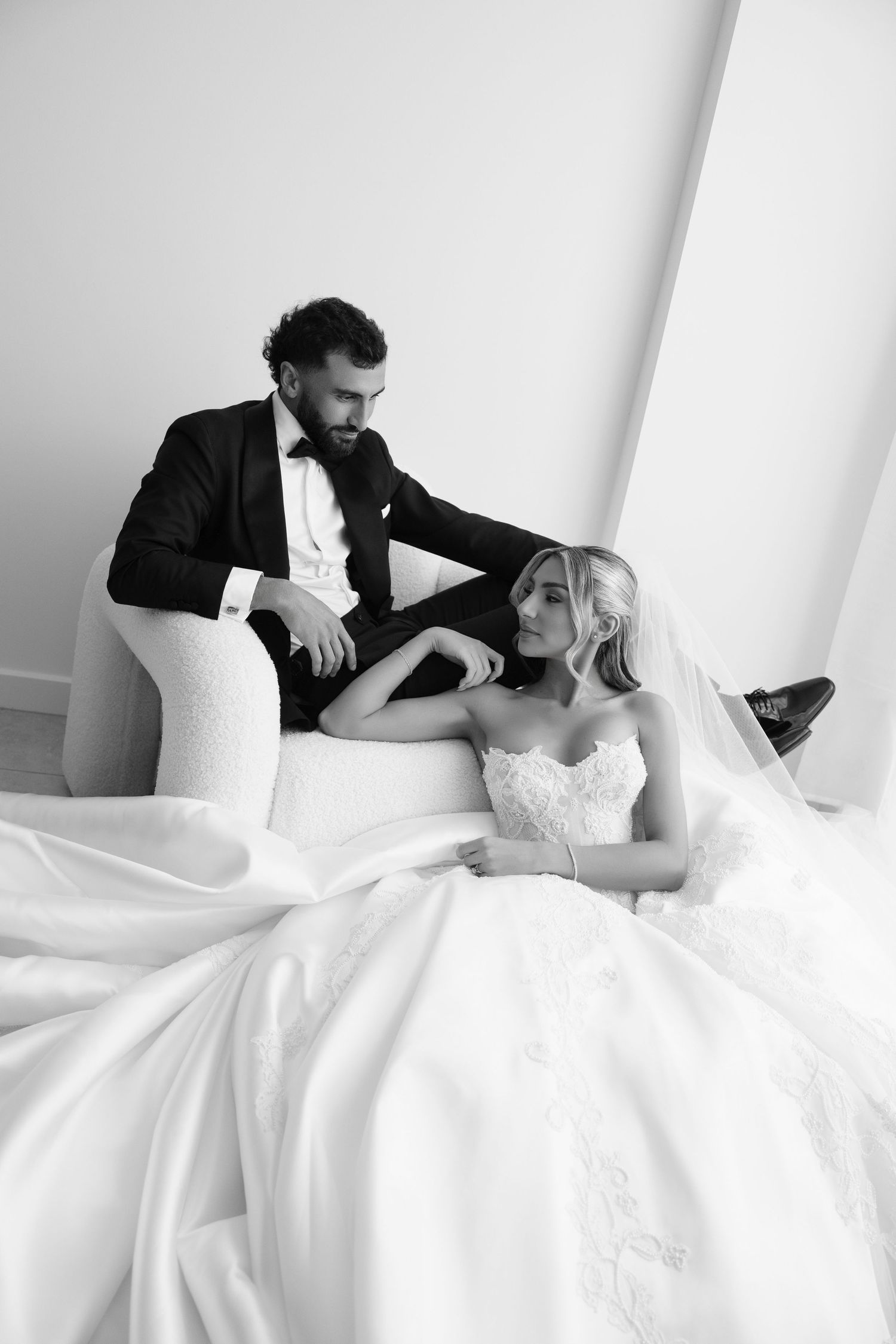 Black and white wedding photo of bride in ballgown and groom in tuxedo posing elegantly on a white sofa.