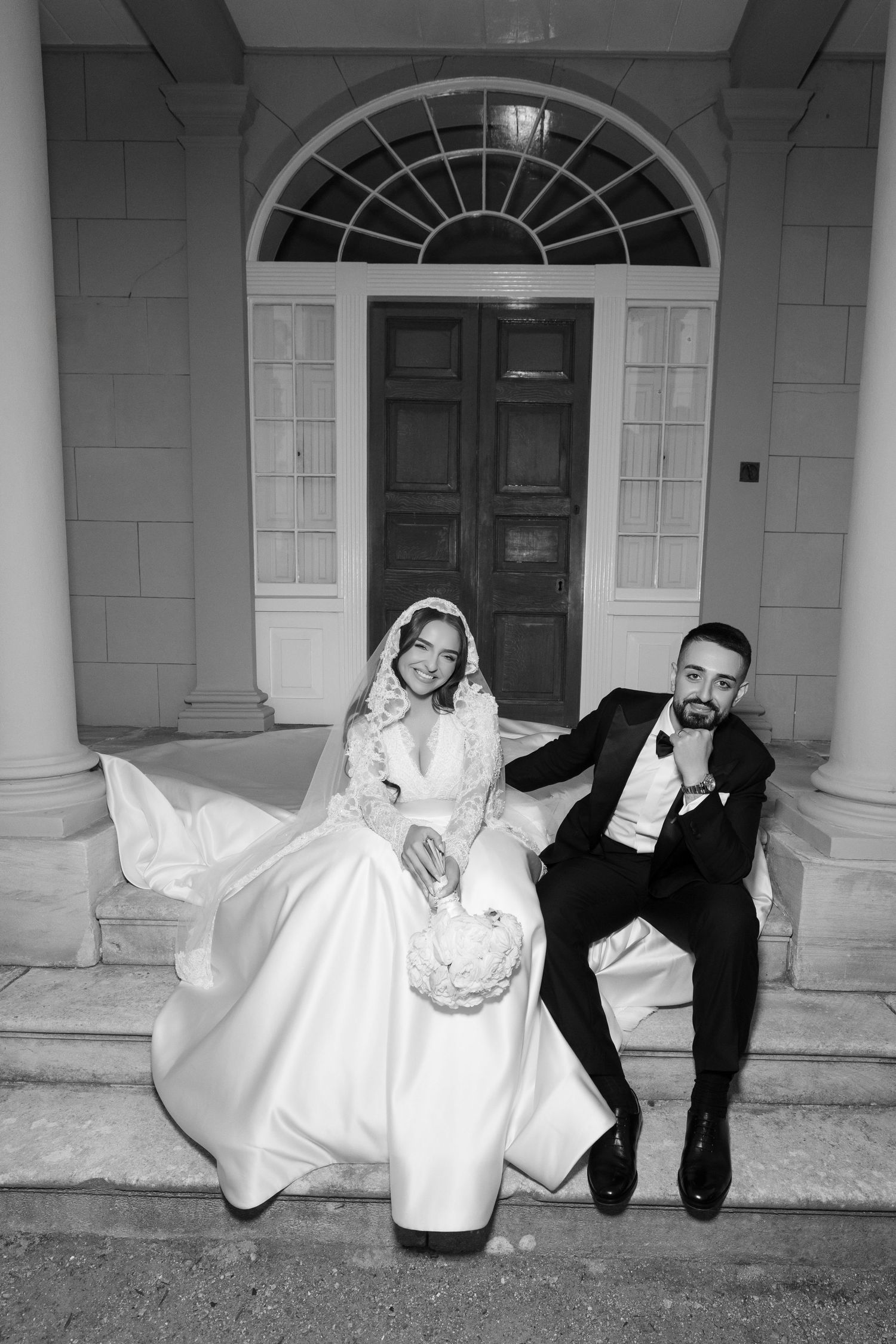Black and white wedding photo of bride in lace gown and groom in tuxedo sitting on steps before elegant doors.
