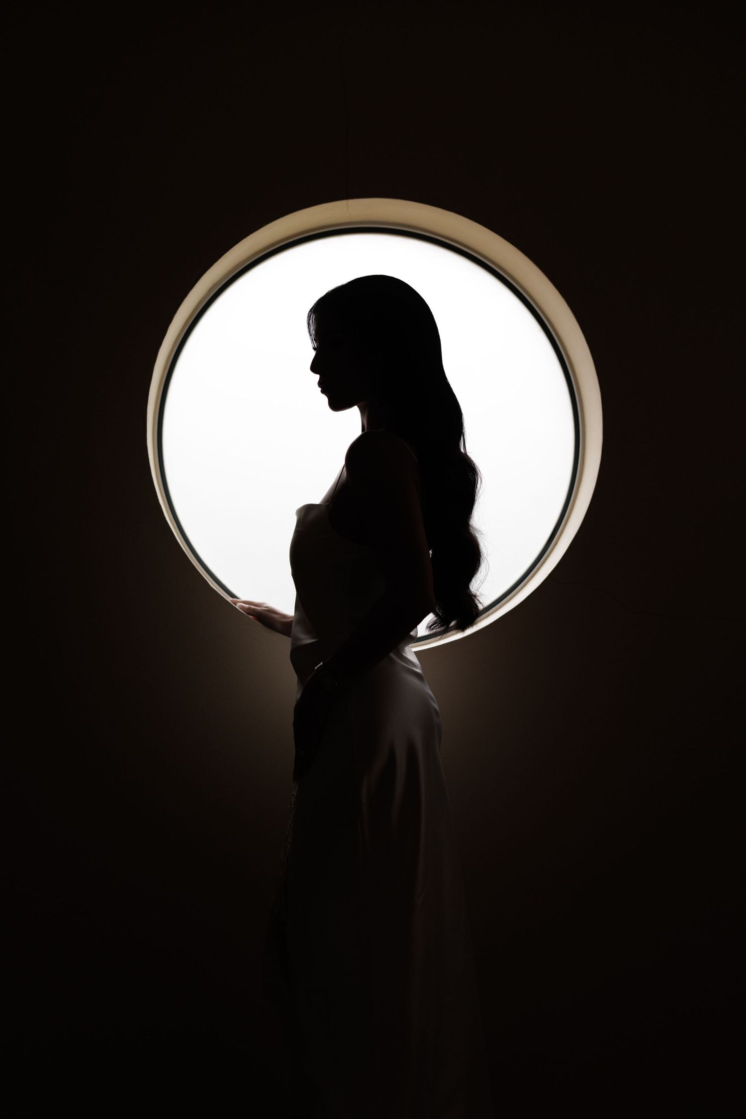 Silhouette of a woman with long hair standing in front of a circular backlit window in a dark room.