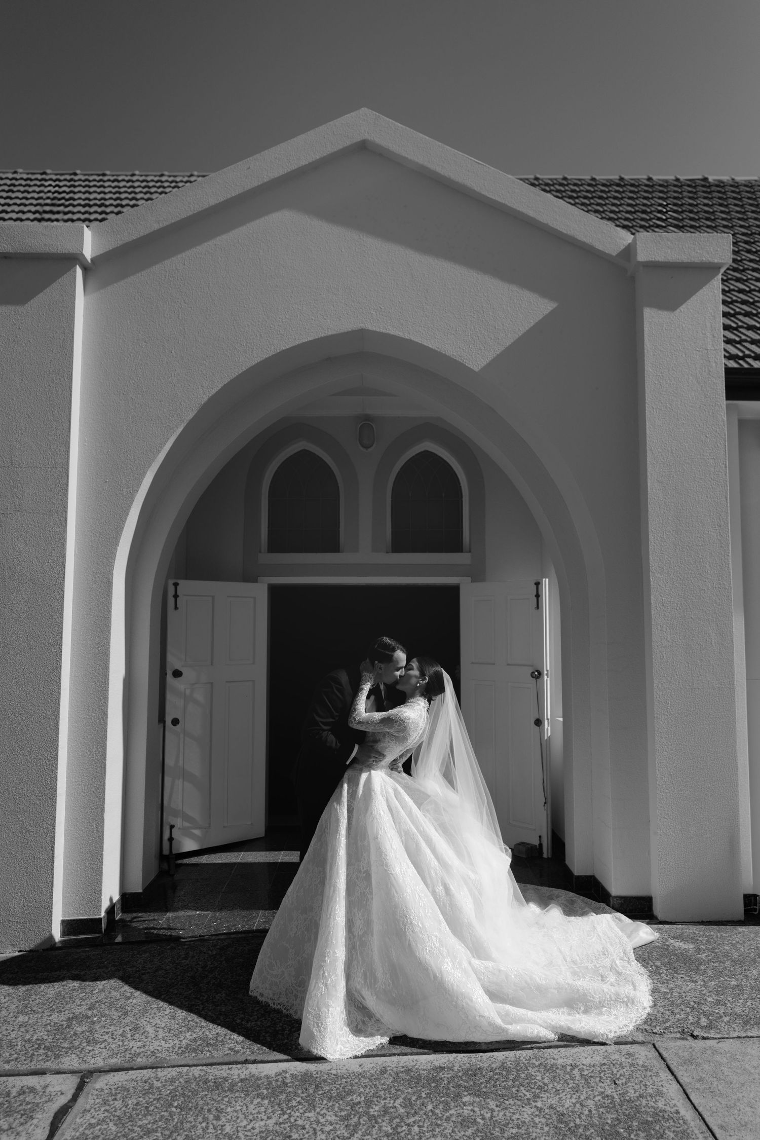 Black and white photo of a bride in a flowing gown standing in a church archway doorway on her wedding day.
