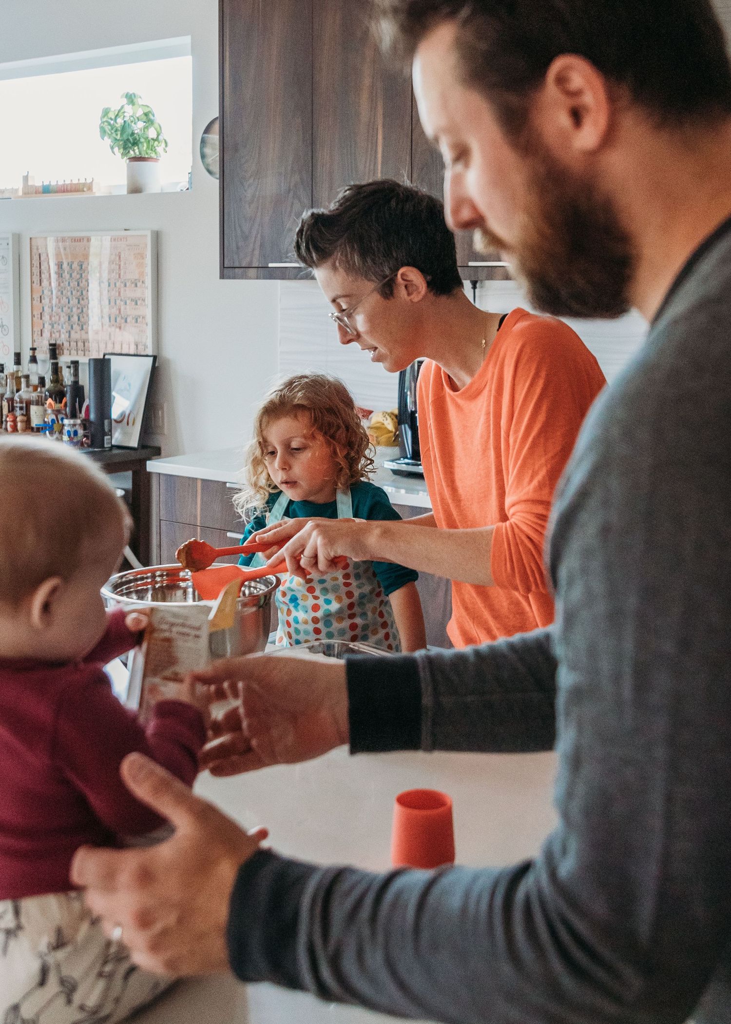 People gather in a kitchen as adults help children with breakfast preparations at the counter during a casual morning moment.