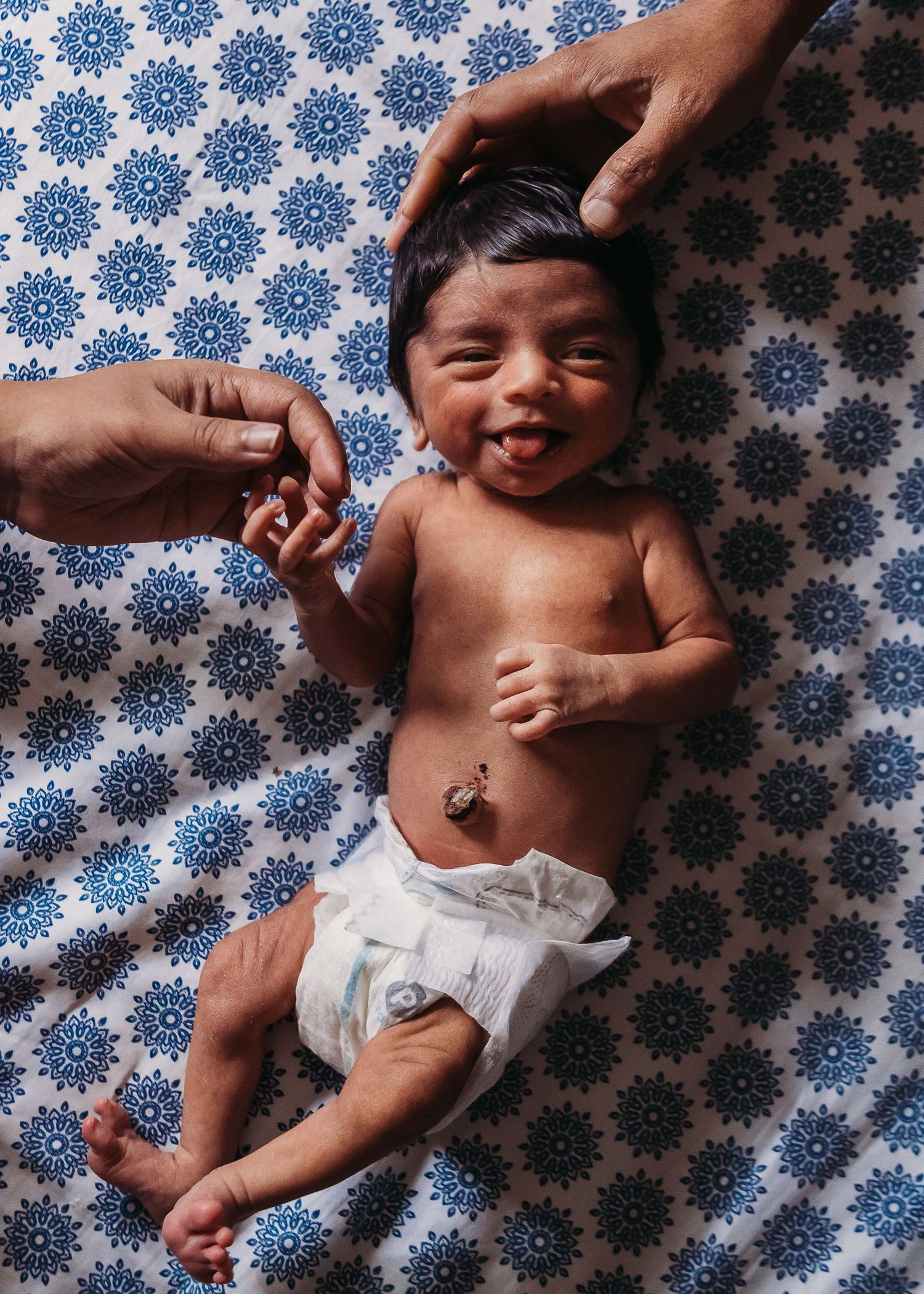 Happy baby wearing a diaper lies on a blue and white patterned blanket while hands reach to tickle and play.