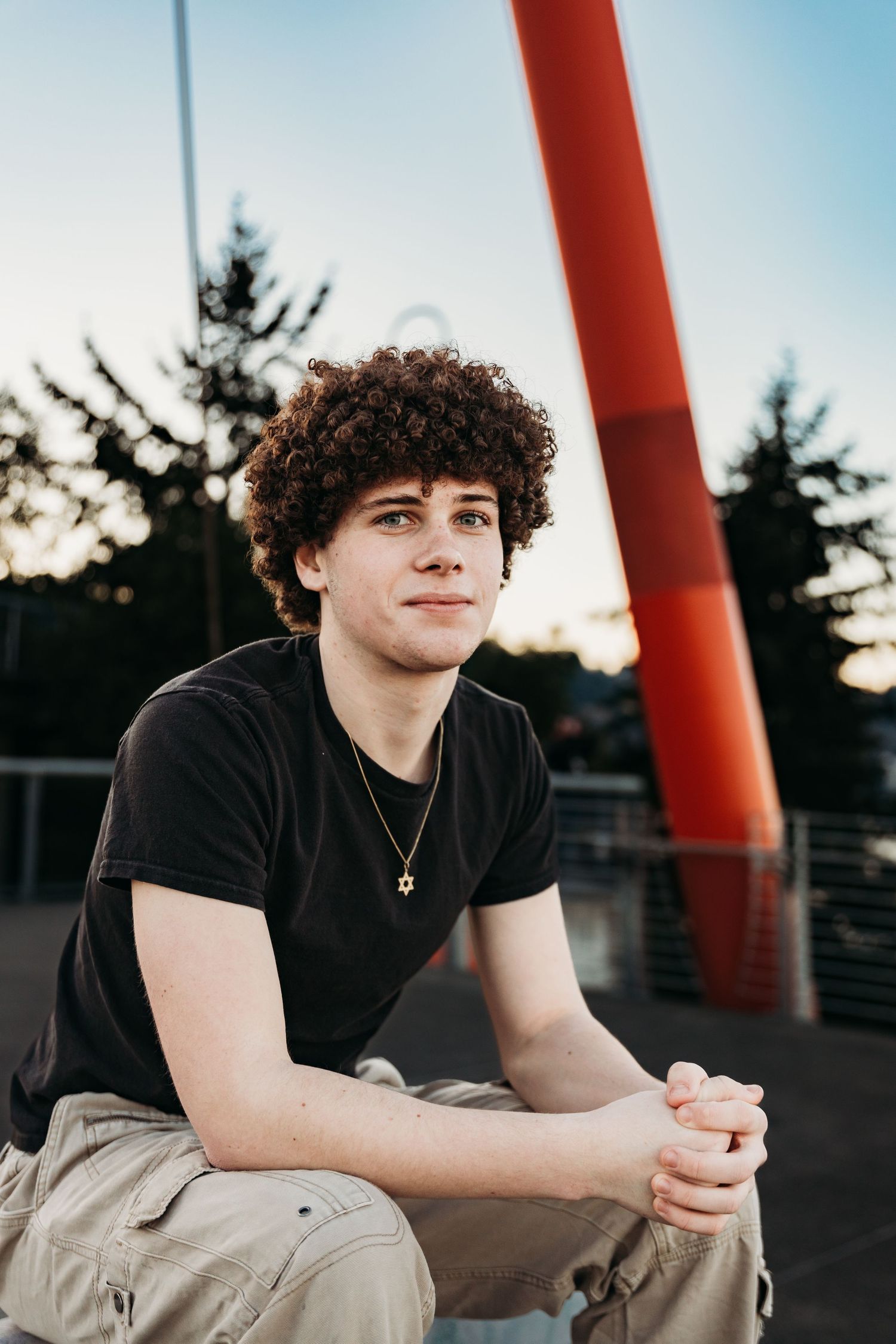 Portrait of a young figure with curly dark hair wearing a black t-shirt sitting outdoors at sunset near a red structural beam.