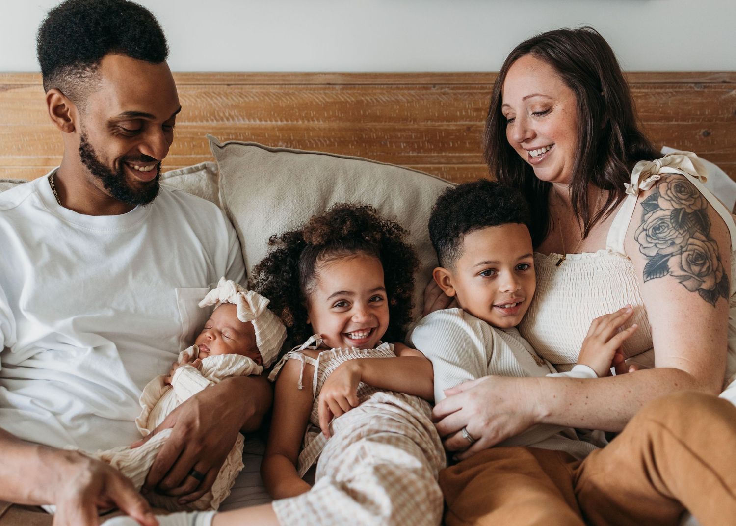 A family lies together relaxing and smiling on a bed with white linens in a warm, candid moment.
