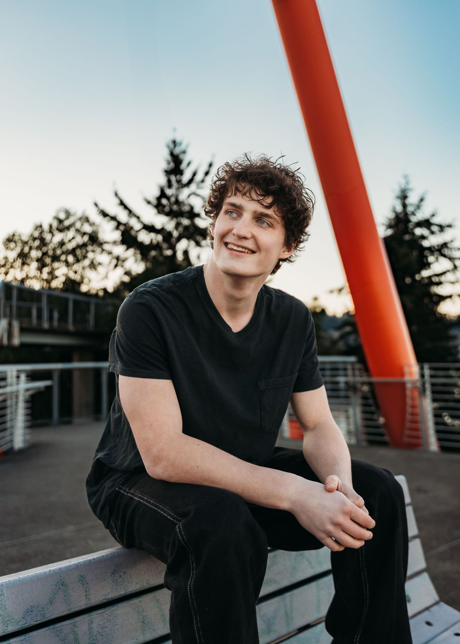 Person sitting on urban bridge at sunset with large red architectural beam in background against blue sky.