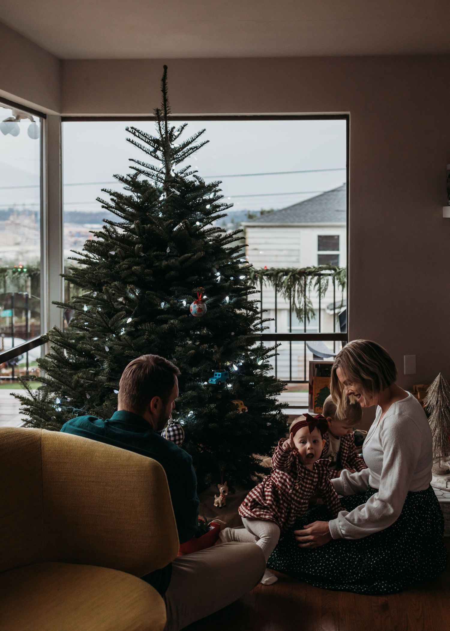 A family sits together by a decorated Christmas tree in their cozy living room near large windows with winter views.
