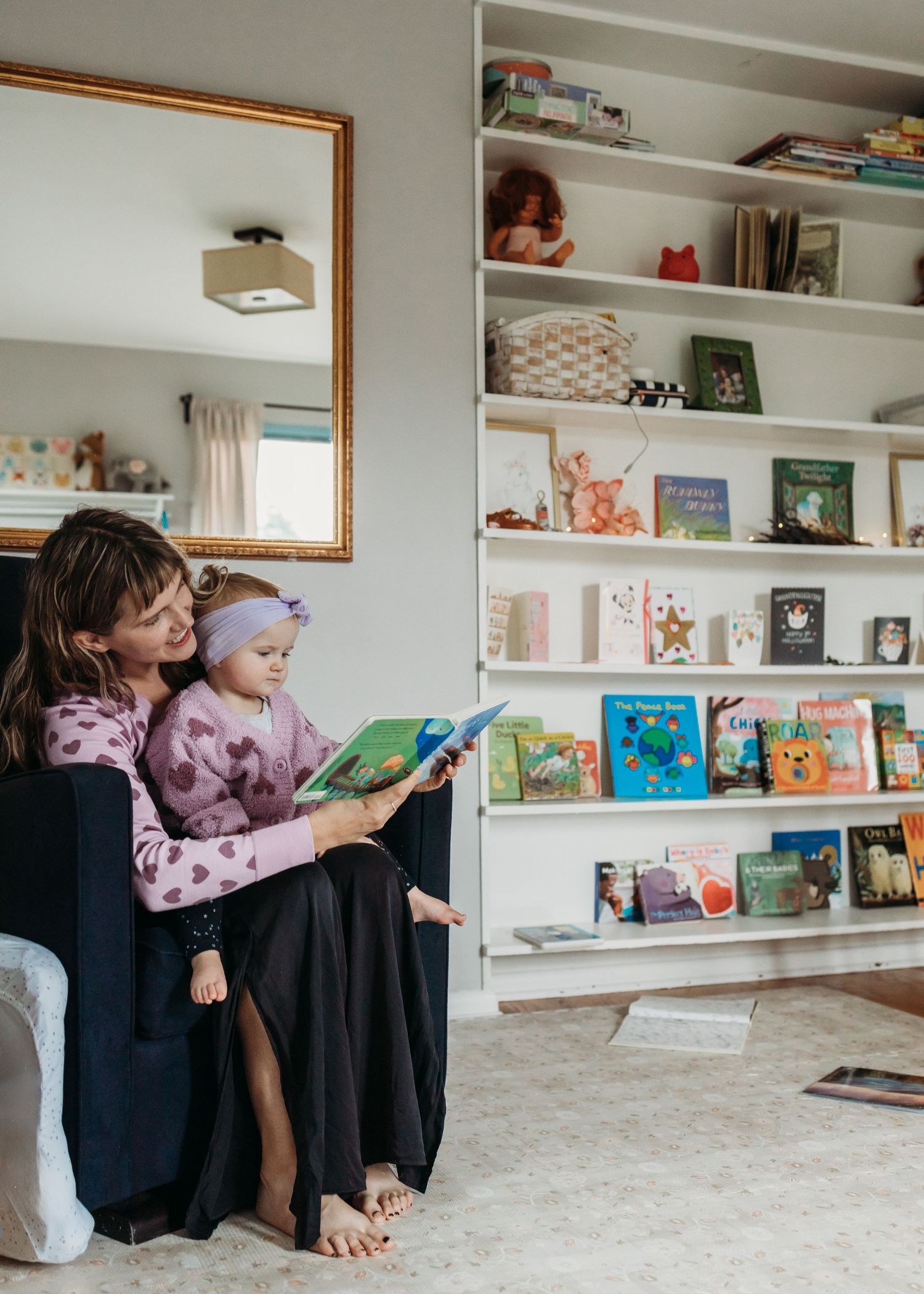 A parent and child sit reading children's books together in a cozy armchair near white shelves filled with colorful books.