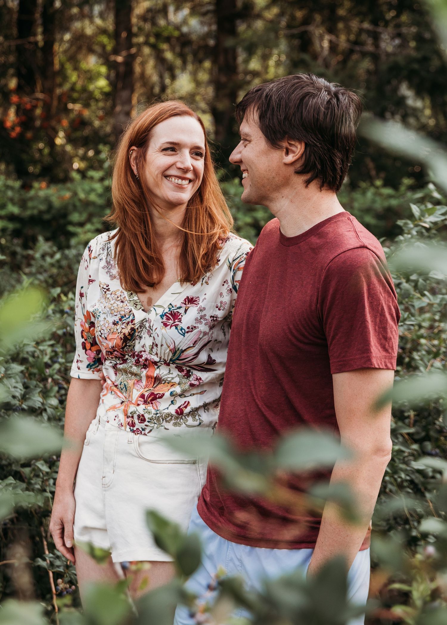 A couple shares a candid moment of laughter while standing together in a lush green forest setting.