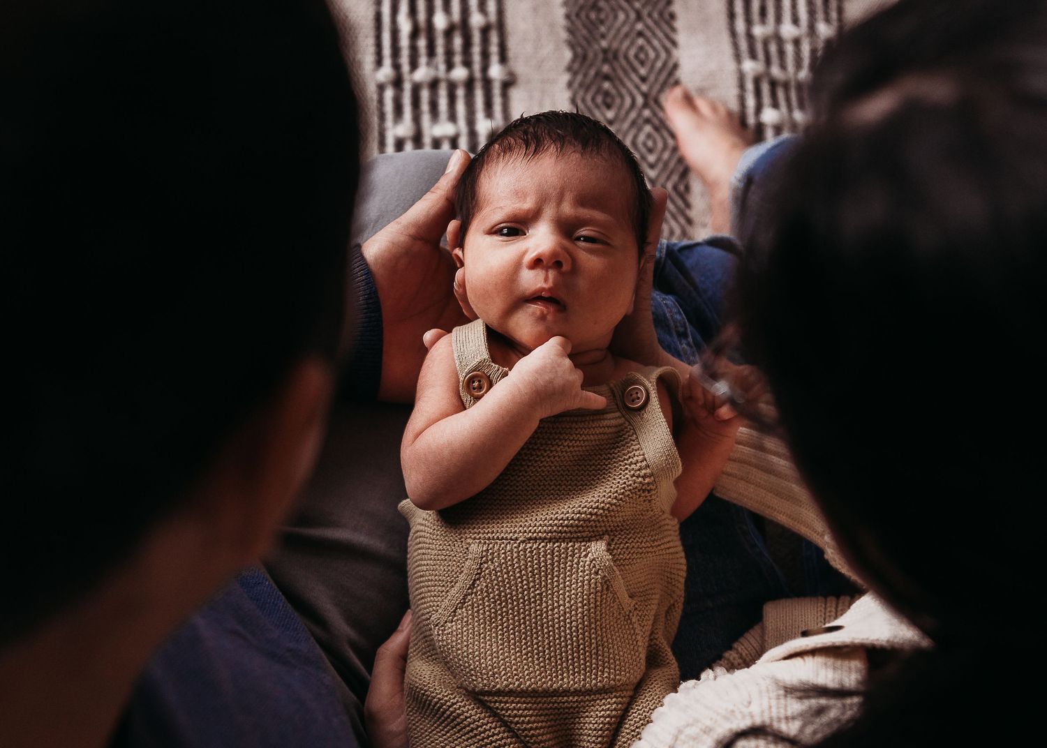 Small child in brown overalls yawns while being held by adults in dim interior setting.