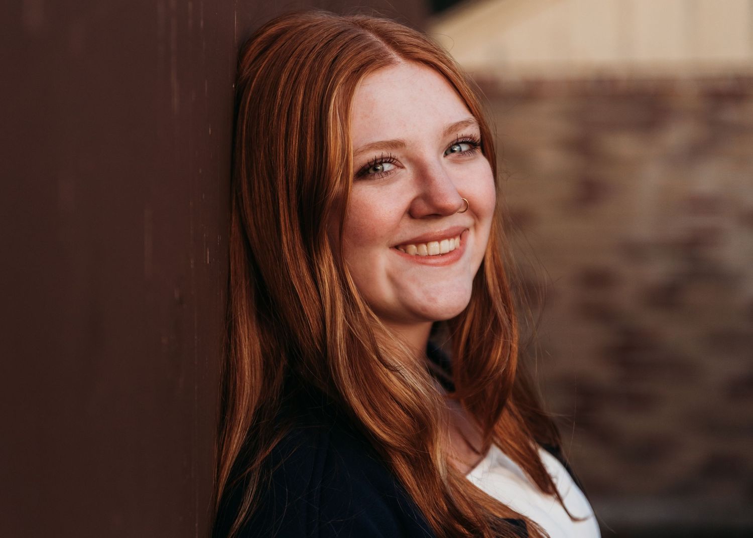 A person with long red hair and a black sweater smiles warmly against a dark textured wall.
