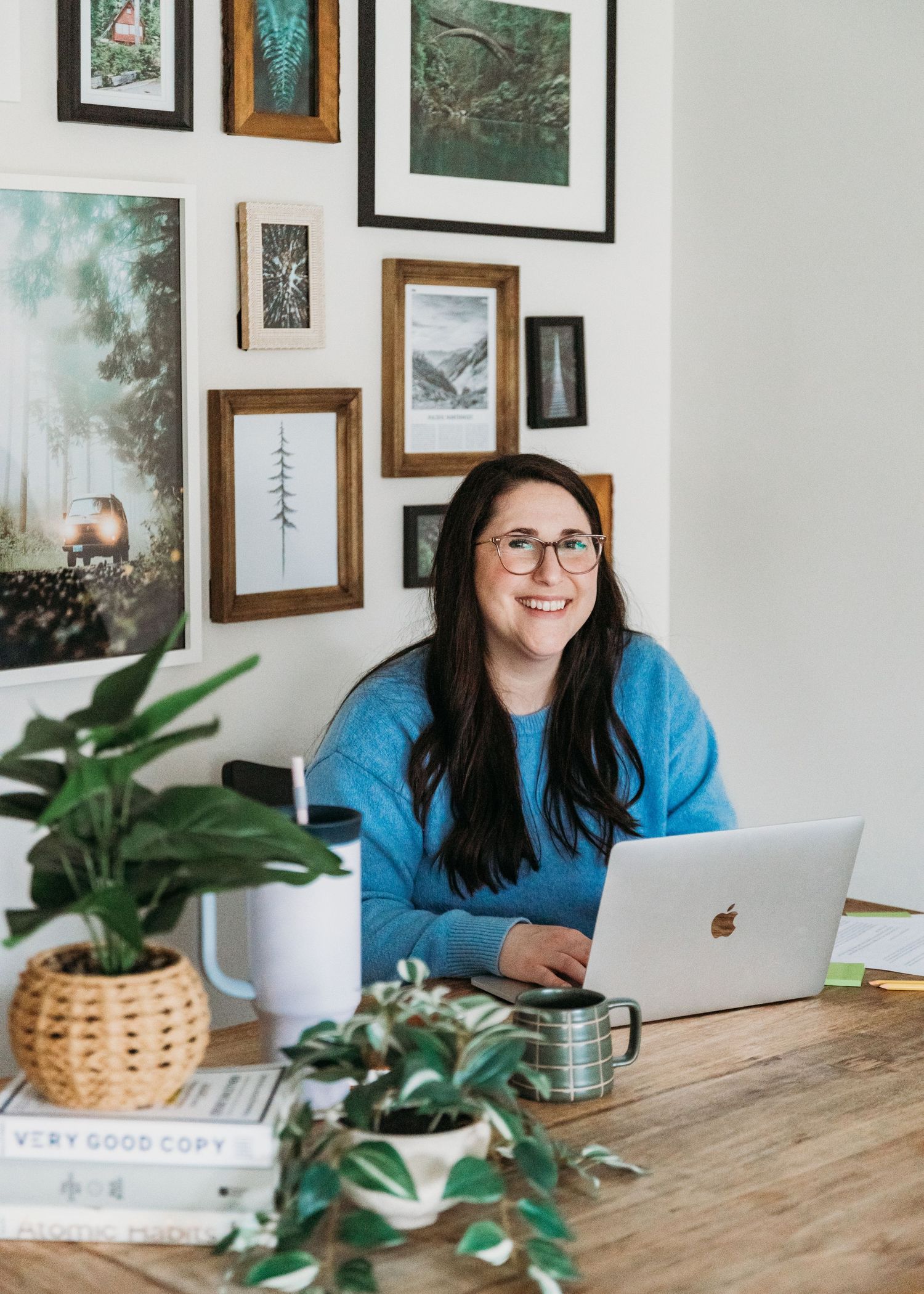 A person in a blue sweater works on a MacBook at a wooden desk decorated with plants and framed wall art.