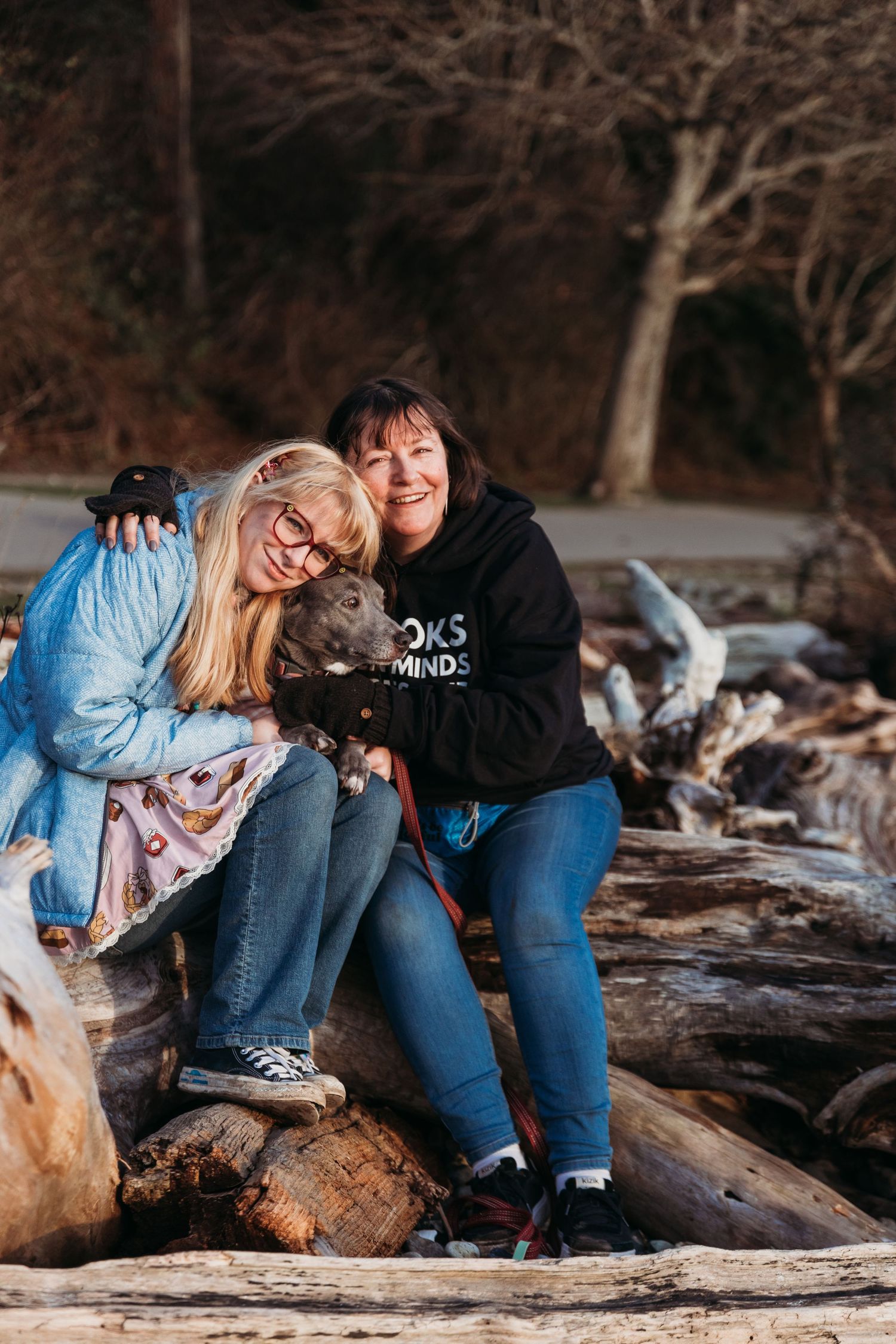 Two people sit on driftwood at the beach with their black dog, sharing laughter at sunset.