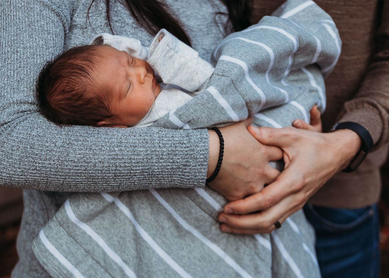 A newborn baby wearing a gray and white striped outfit is held gently in caring arms.