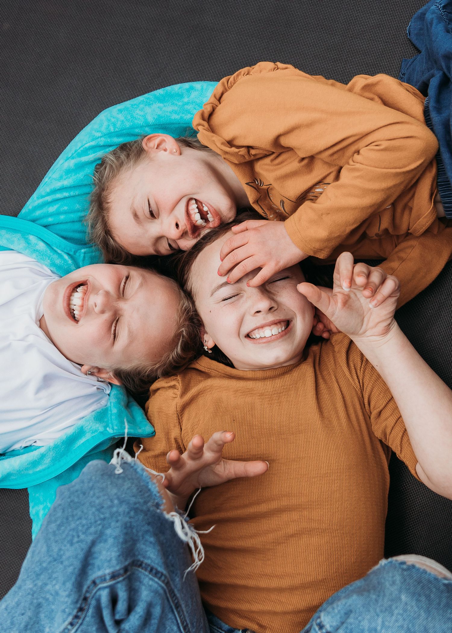 Three children lie together on the floor laughing joyfully against a dark background with teal and brown accents.