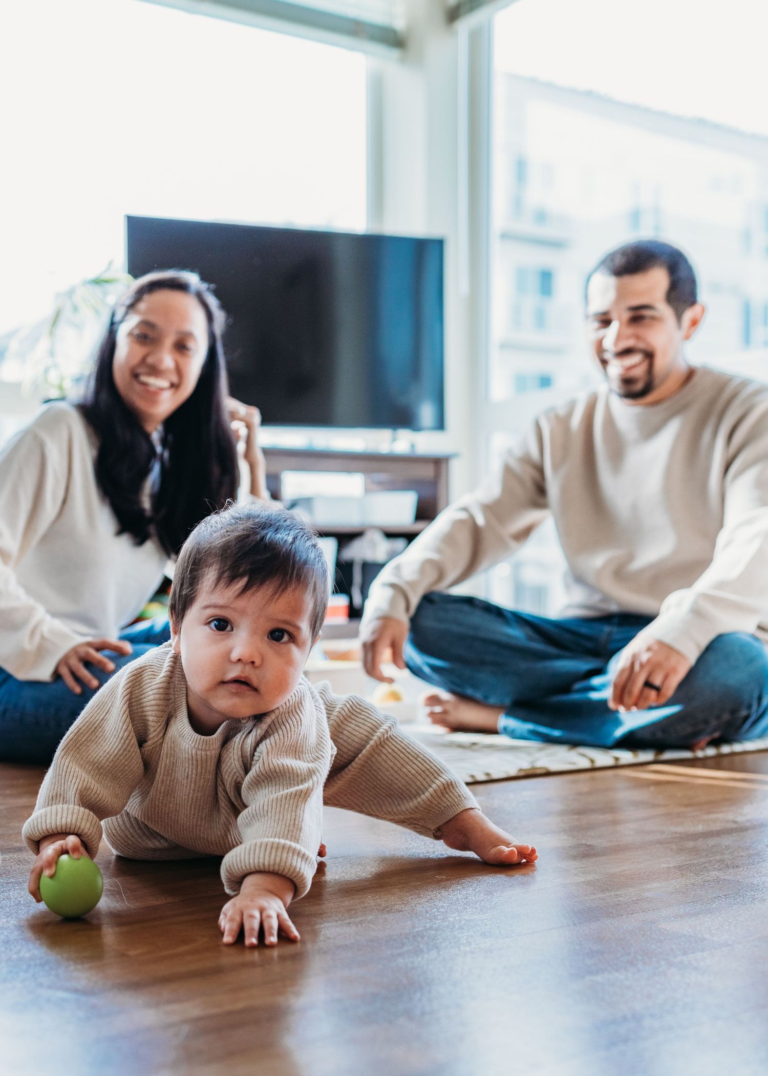Family relaxing together on hardwood floor with baby crawling and holding green toy in modern living room.