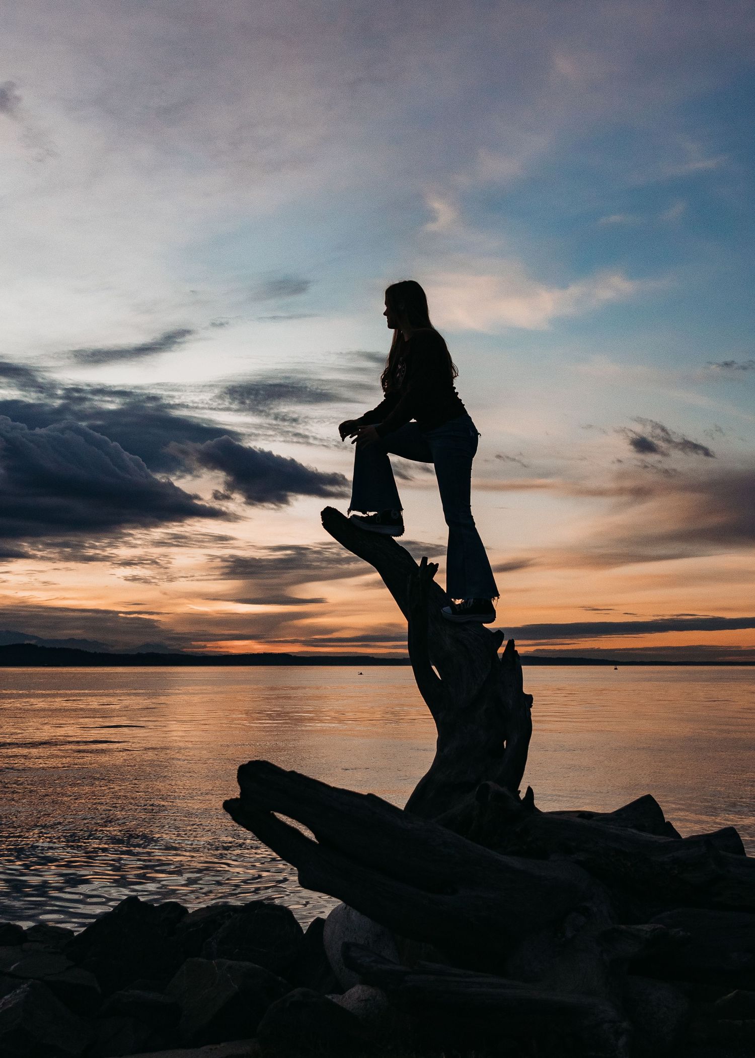 Silhouette of person standing on driftwood log against dramatic sunset sky over calm water at dusk.