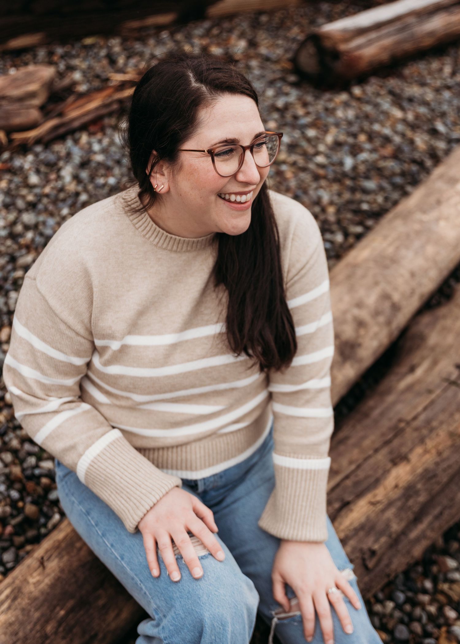 Someone in glasses and a cream striped sweater sits casually on wooden logs while laughing outdoors.