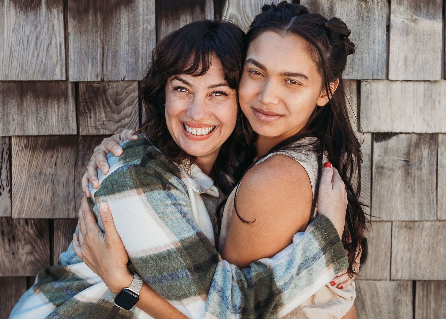 Two friends wrapped in a plaid blanket share a joyful moment while leaning against a wooden wall.