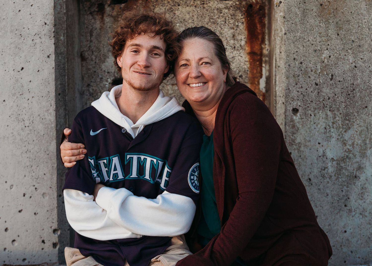 Two people share a warm moment while one wears a vintage Seattle Mariners sweatshirt against a grey concrete wall.