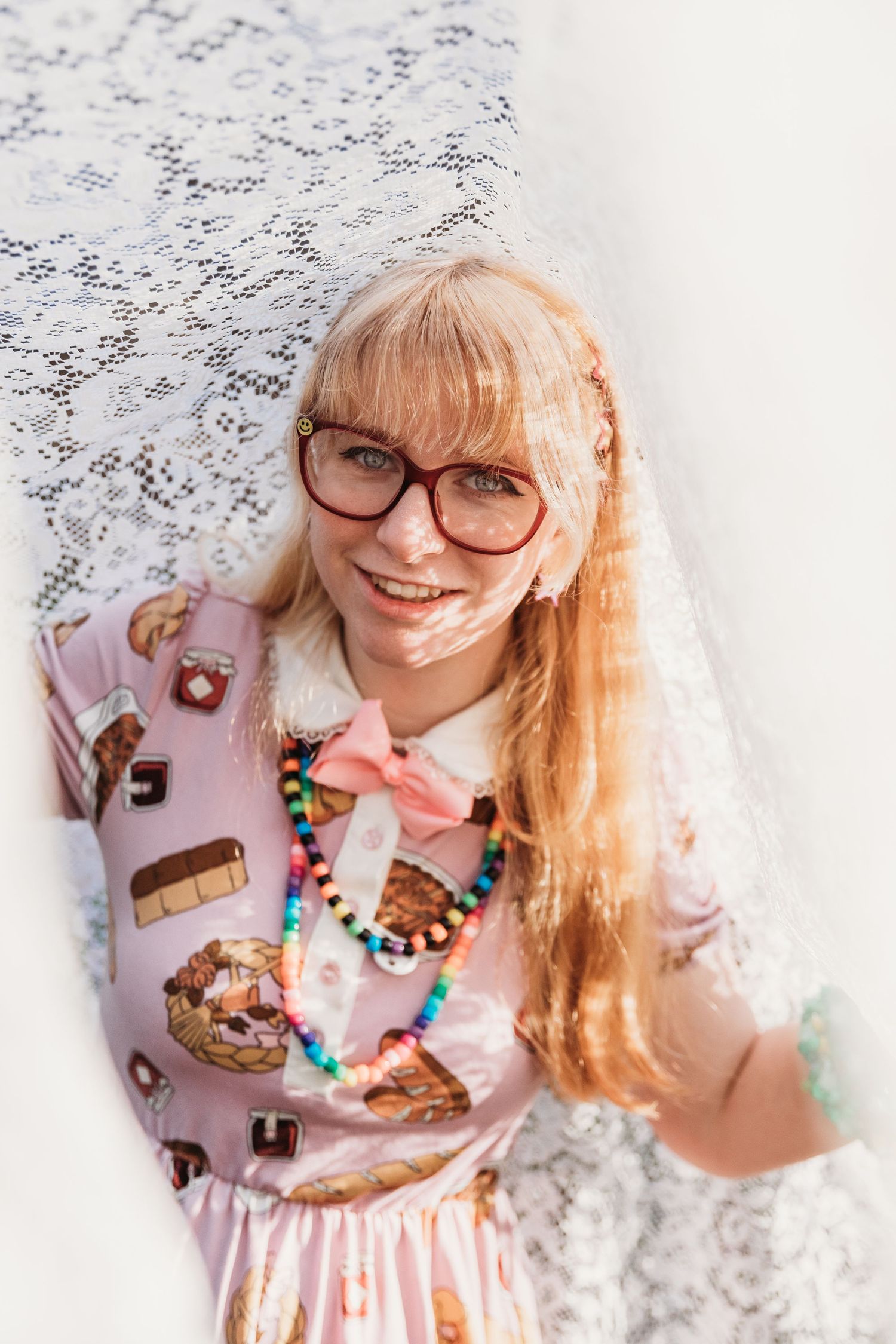A person wearing glasses and a pink dress with food prints smiles against a white background.