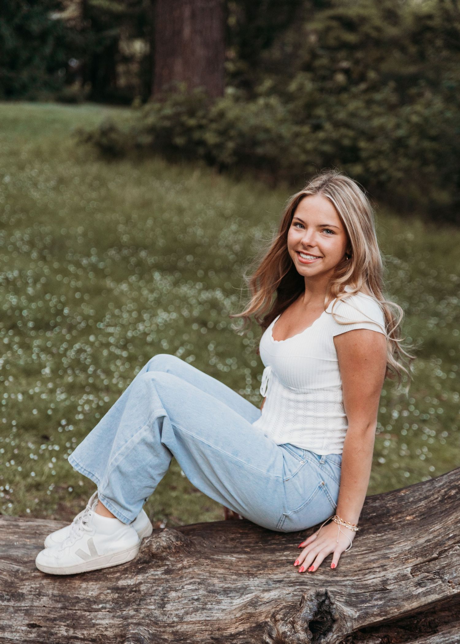 Someone in a white top and light jeans sits casually on a fallen log in a grassy outdoor setting on a summer day.