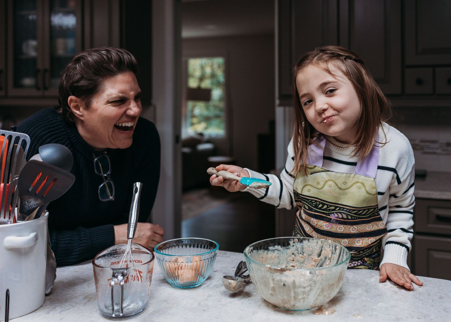 Two people laugh while baking together in a kitchen with mixing bowls and ingredients on the counter.