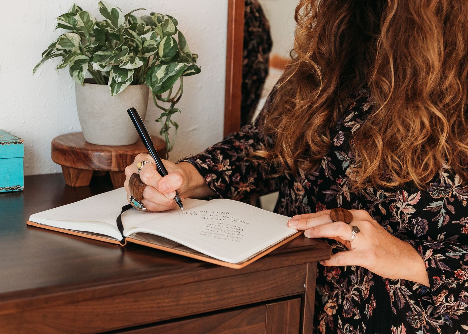 Person with long wavy hair writing in a journal at a wooden desk with a houseplant nearby.